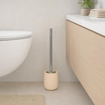 Eine beige WC-Bürste steht neben der Toilette im hellen Bad.