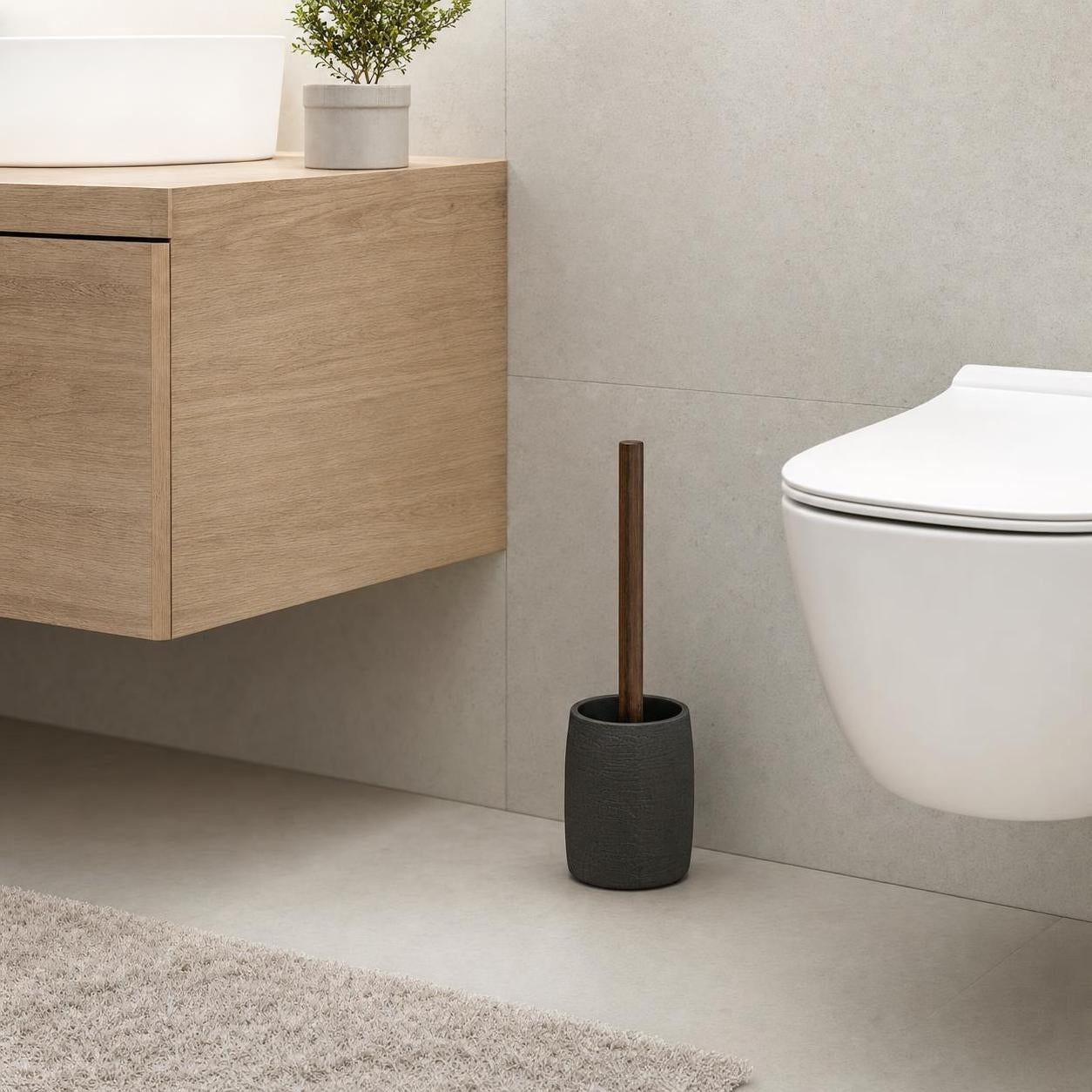 Eine WC-Bürste mit Holzgriff steht im grauen Halter neben der Toilette.