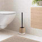 Eine beige Toilettenbürste mit schwarzem Griff steht neben dem WC.