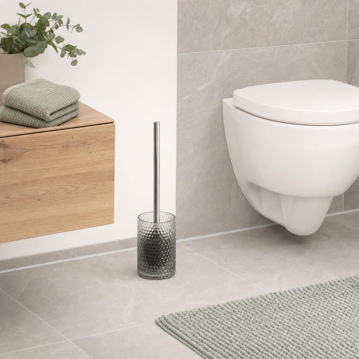 Eine Toilettenbürste im Glasbehälter steht neben dem WC.
