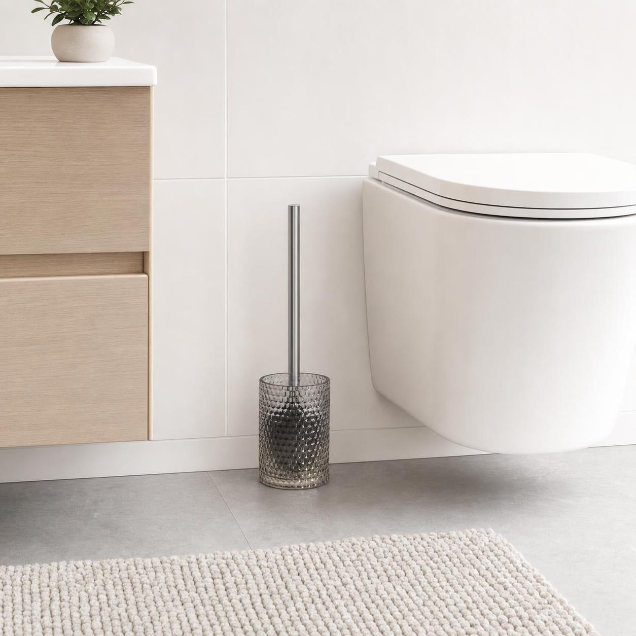 Eine Toilettenbürste im grauen Glasbehälter steht neben der Toilette.
