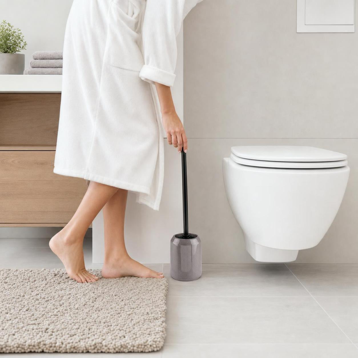 Eine graue WC-Bürste steht neben der Toilette im hellen Bad.