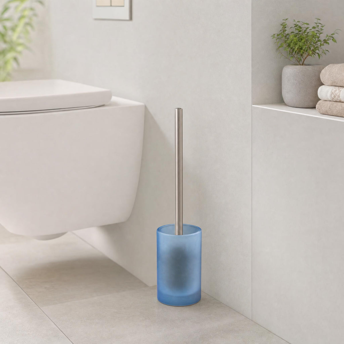Blaue WC-Bürste aus Glas in hellem Bad neben Wand-WC und Handtüchern.