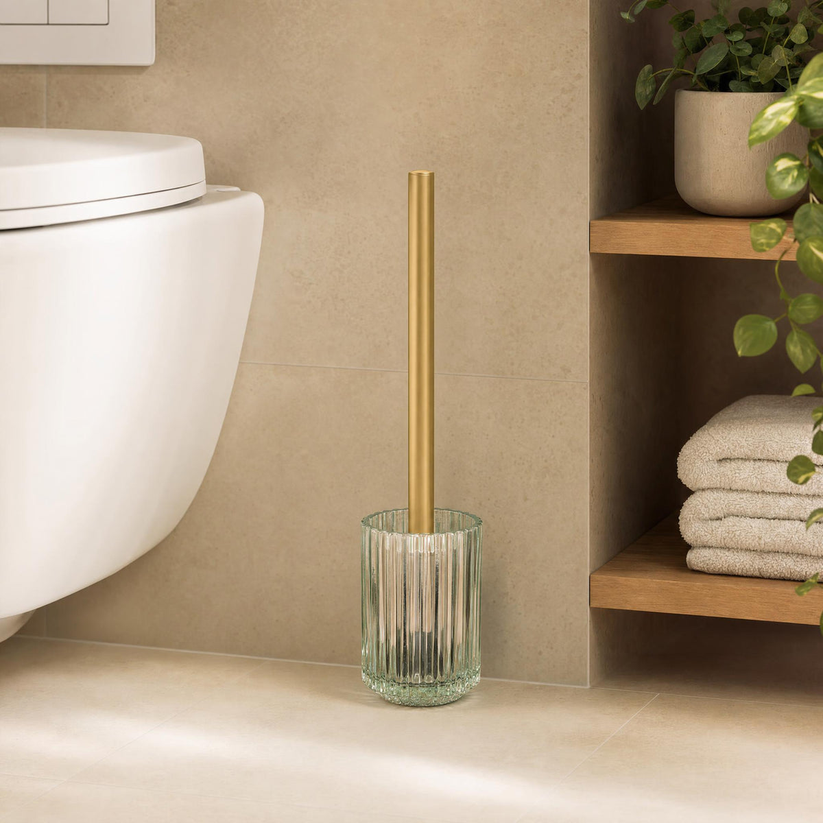 WC-Bürste im Glasbehälter mit goldfarbenem Griff neben Wand-WC und Holzregal.