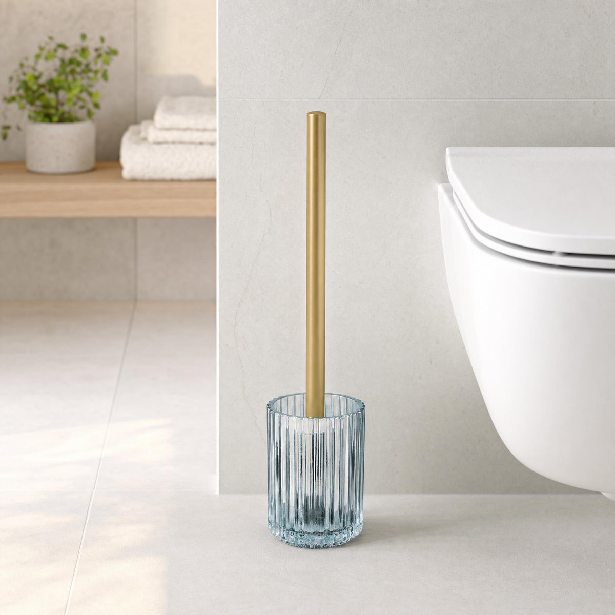 WC-Bürste mit goldenem Griff im Glasbehälter neben wandhängendem WC.