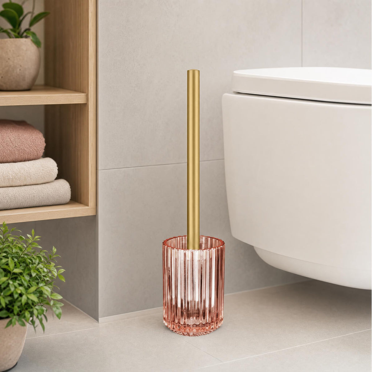 Rosa WC-Bürste aus Glas mit goldfarbenem Handgriff neben modernem Hänge-WC.