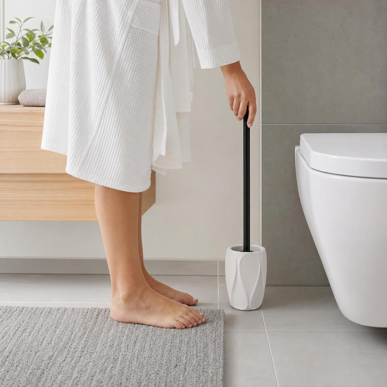 Eine WC-Bürste mit schwarzem Griff steht im weißen Halter neben der Toilette.