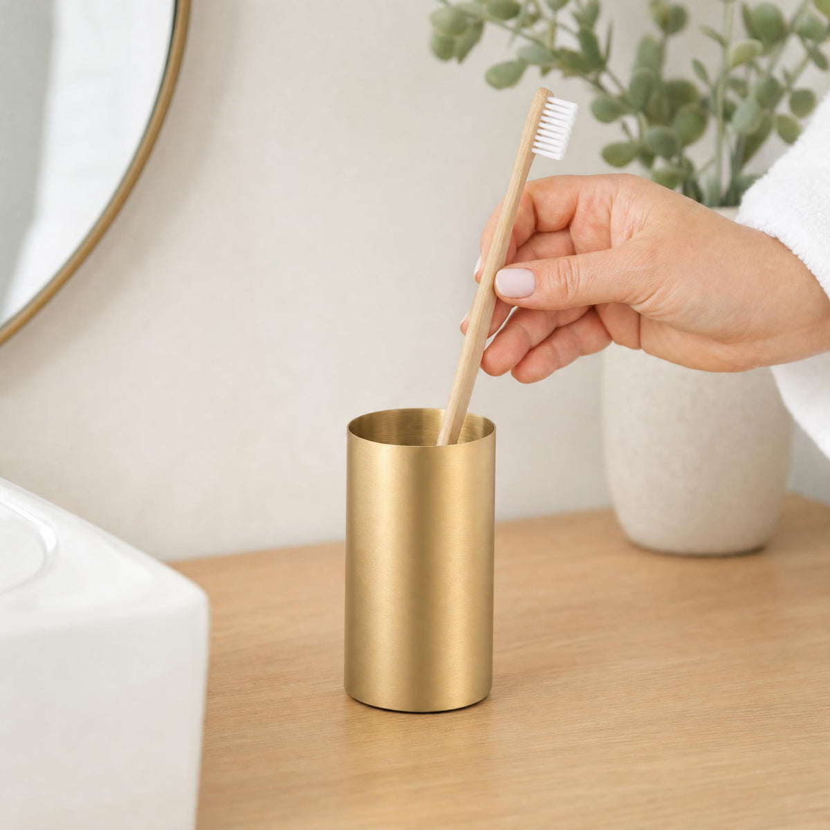 Hand legt Bambuszahnbürste in goldfarbenen Becher auf Badezimmerablage.