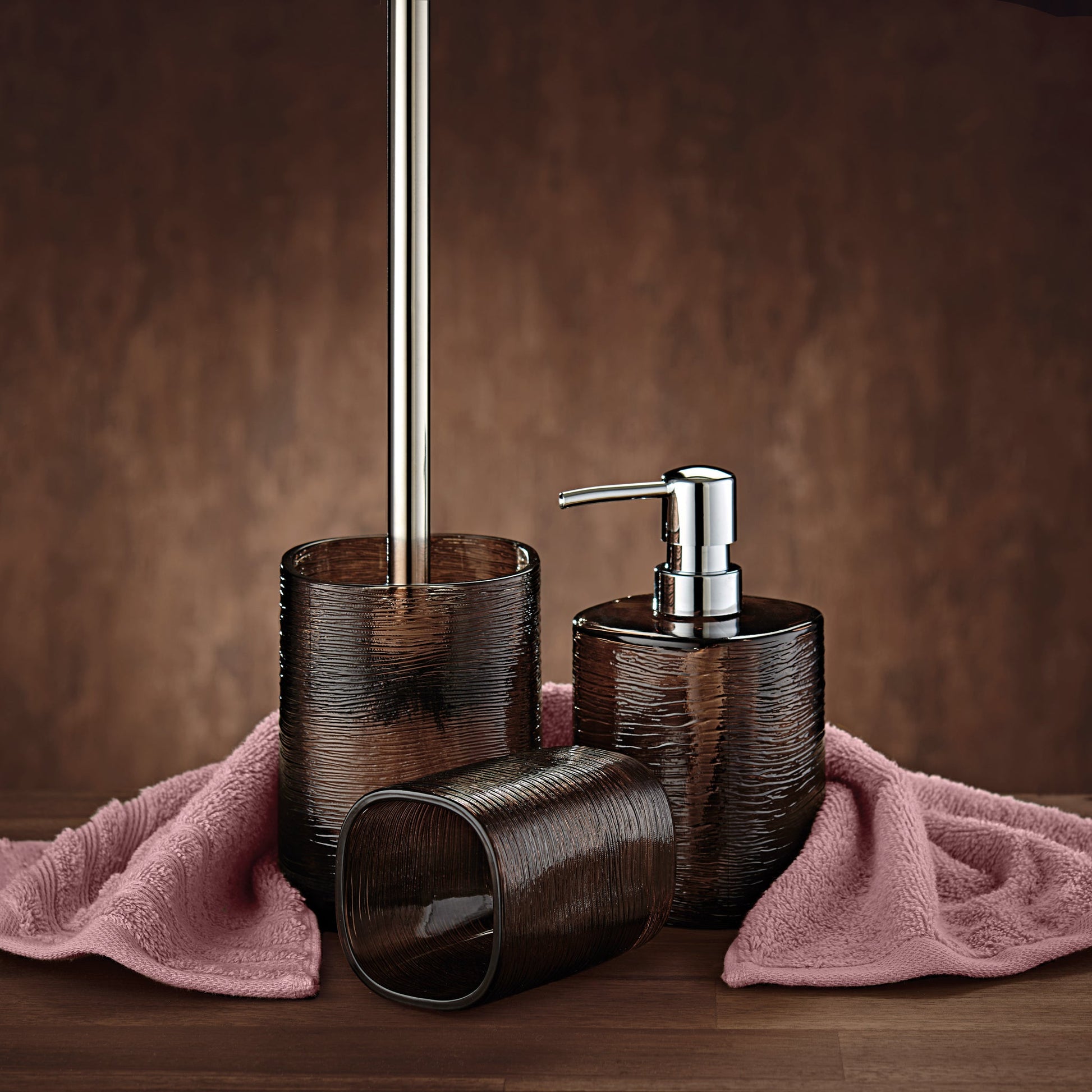 Zahnputzbecher QUBO Nussbraun mit Seifenspender und WC-Bürste auf rosa Handtuch im Bad.