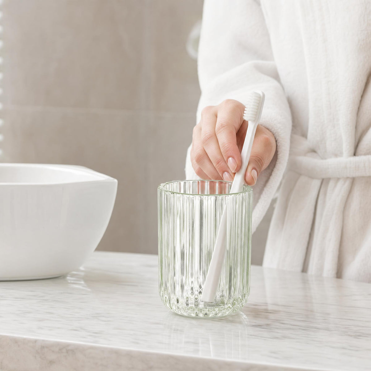 Hand mit Zahnbürste über geriffeltem Glasbecher auf hellem Badwaschtisch.