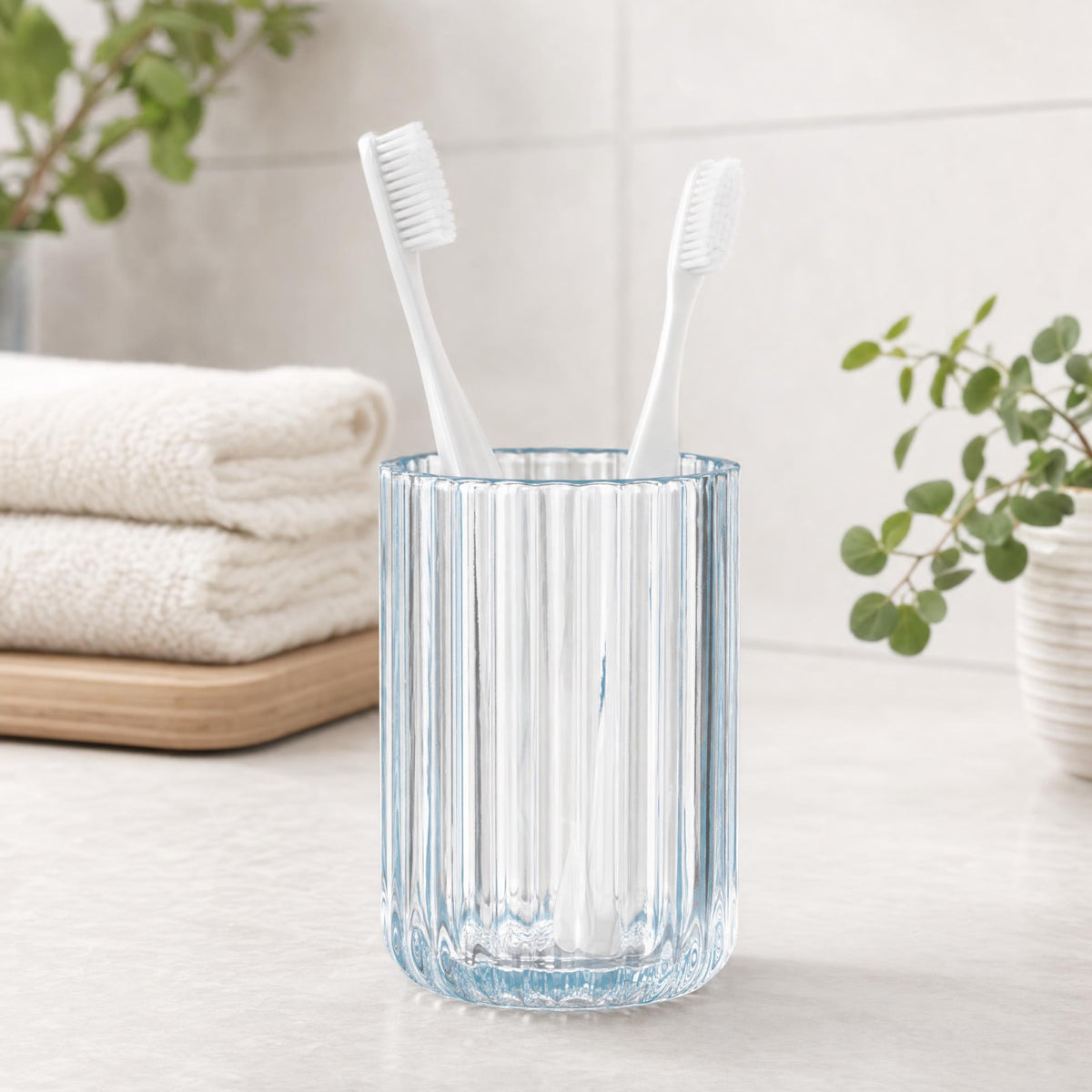 Zwei weiße Zahnbürsten im geriffelten Glasbecher auf hellem Waschtisch.