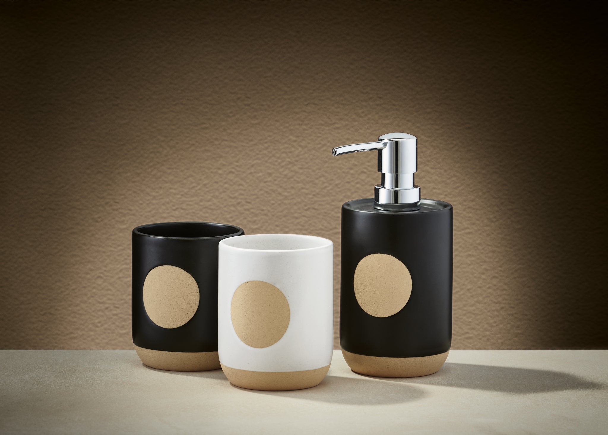 Zahnputzbecher SPOT Weiß steht zwischen zwei schwarzen Bechern und einem Seifenspender auf einer Fläche.