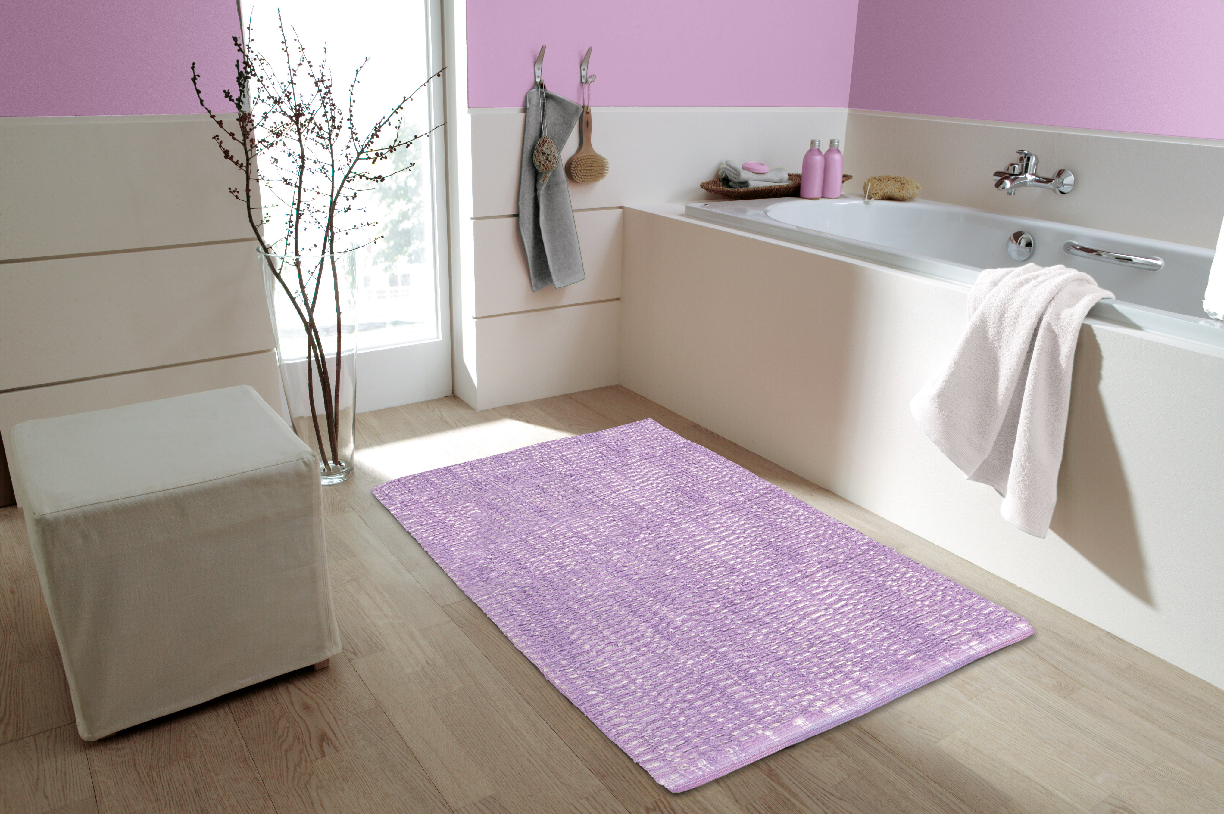 Fliederfarbener Badteppich in modernem Badezimmer mit Badewanne, Hocker und Handtüchern, dekoriert mit Pflanzen und Accessoires.