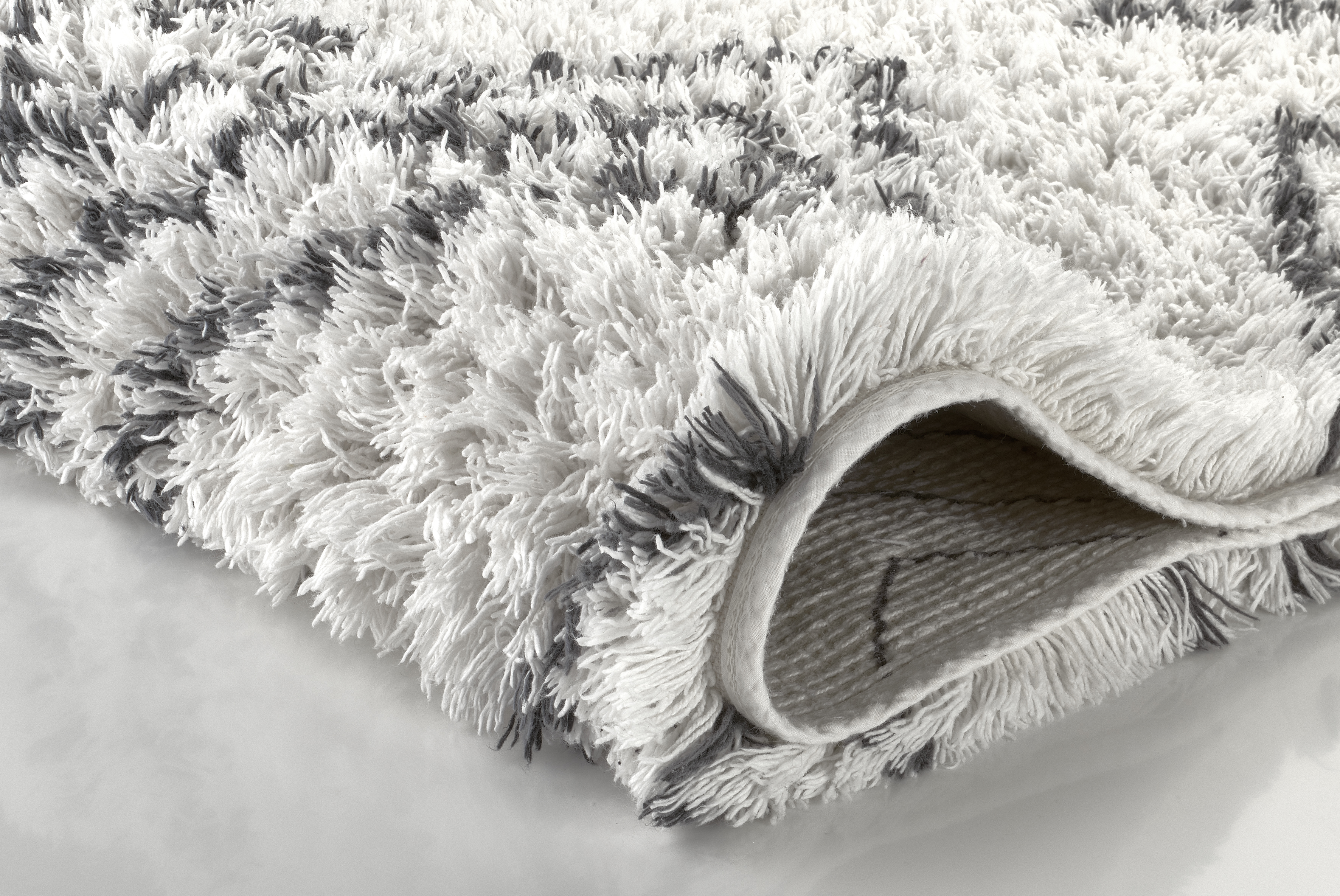 Weißer Hochflor-Teppich mit grauen Akzenten, weiche Textur, eingerollte Ecke zeigt Rückseite. Ideal für gemütliche Wohnräume.