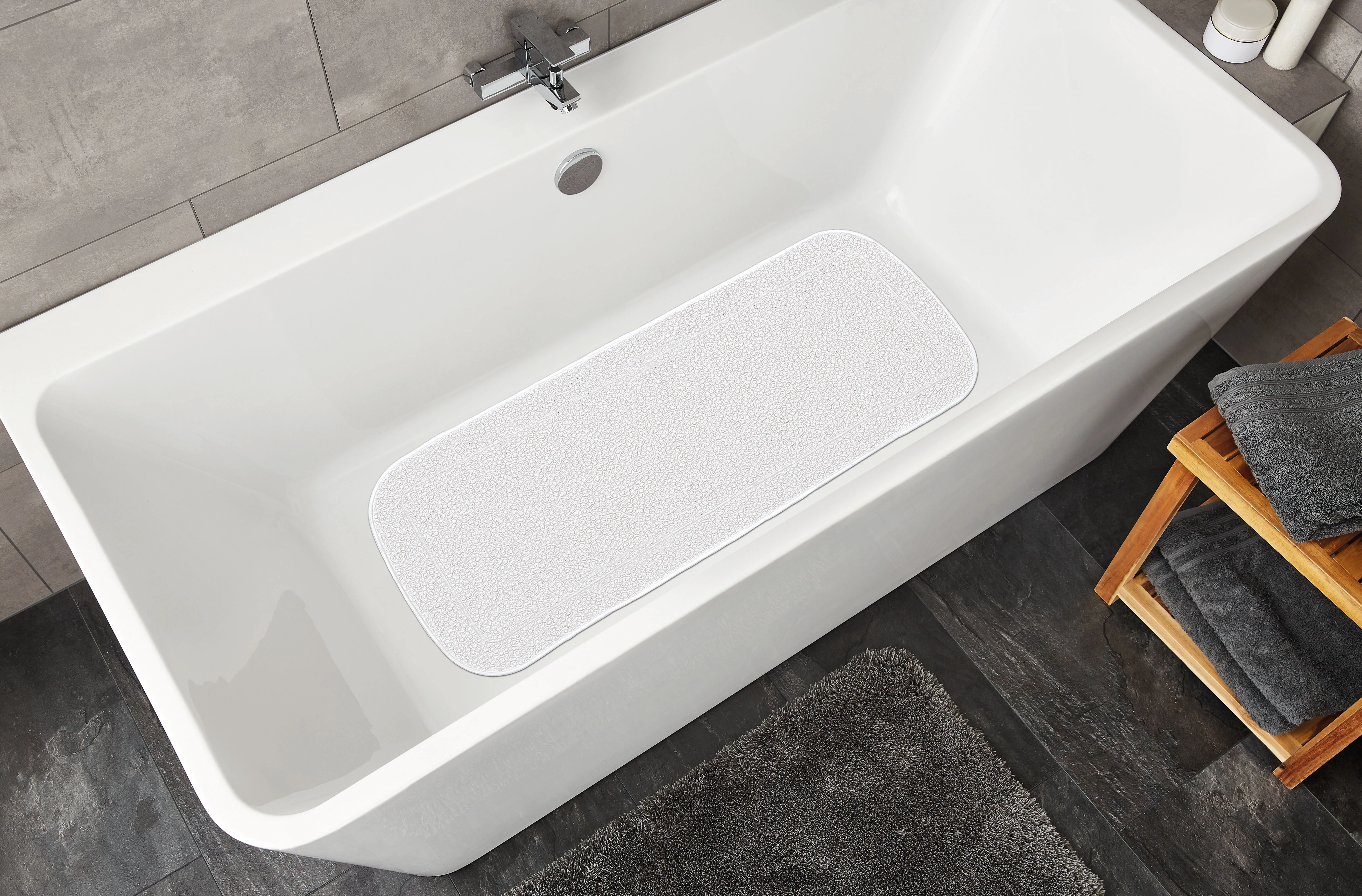 Weiße Badewannenmatte in einer modernen Badewanne, umgeben von grauen Fliesen und Handtüchern.