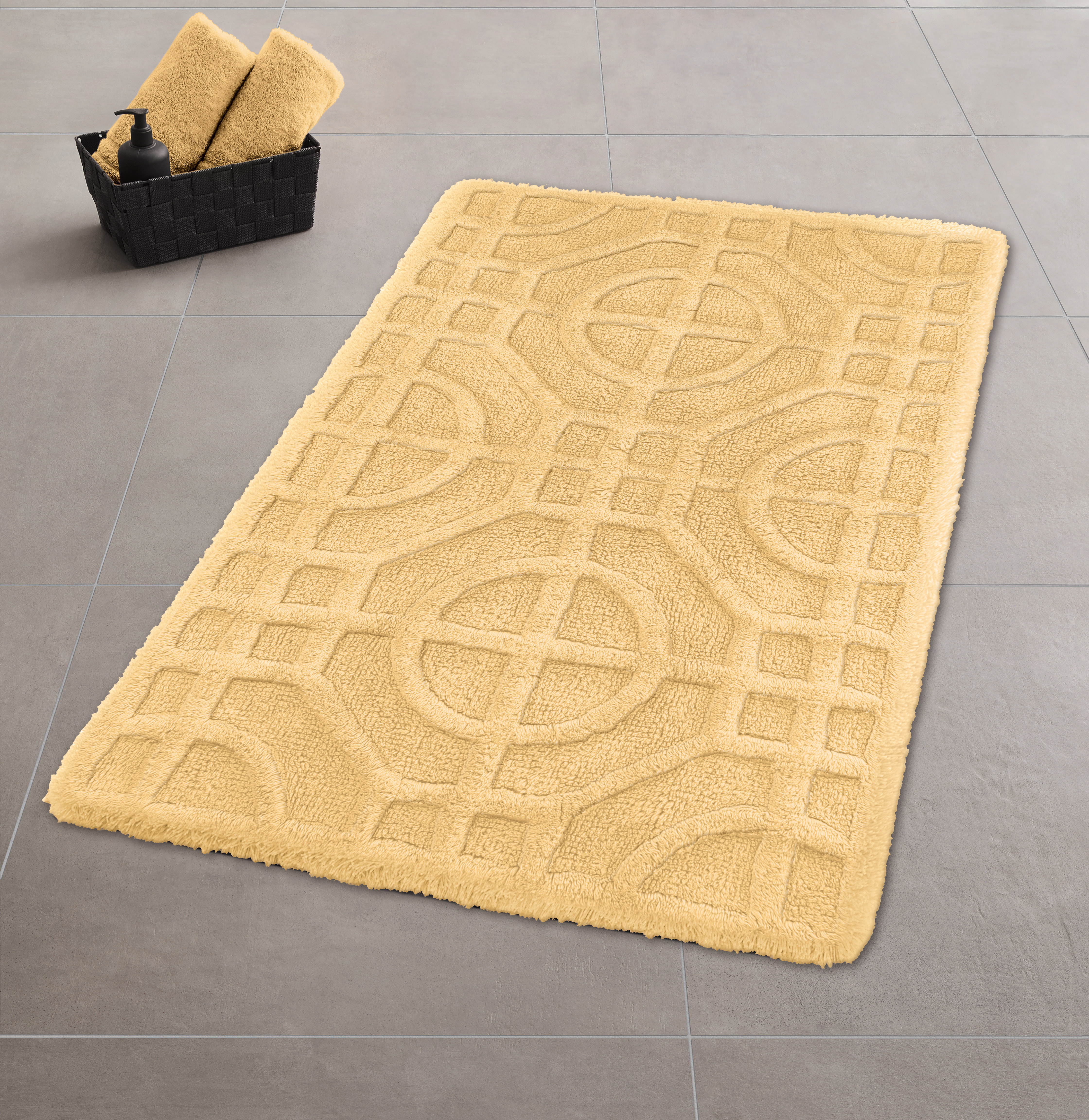 Gelber Badteppich mit geometrischem Muster auf grauem Fliesenboden, daneben ein schwarzer Korb mit Handtüchern.