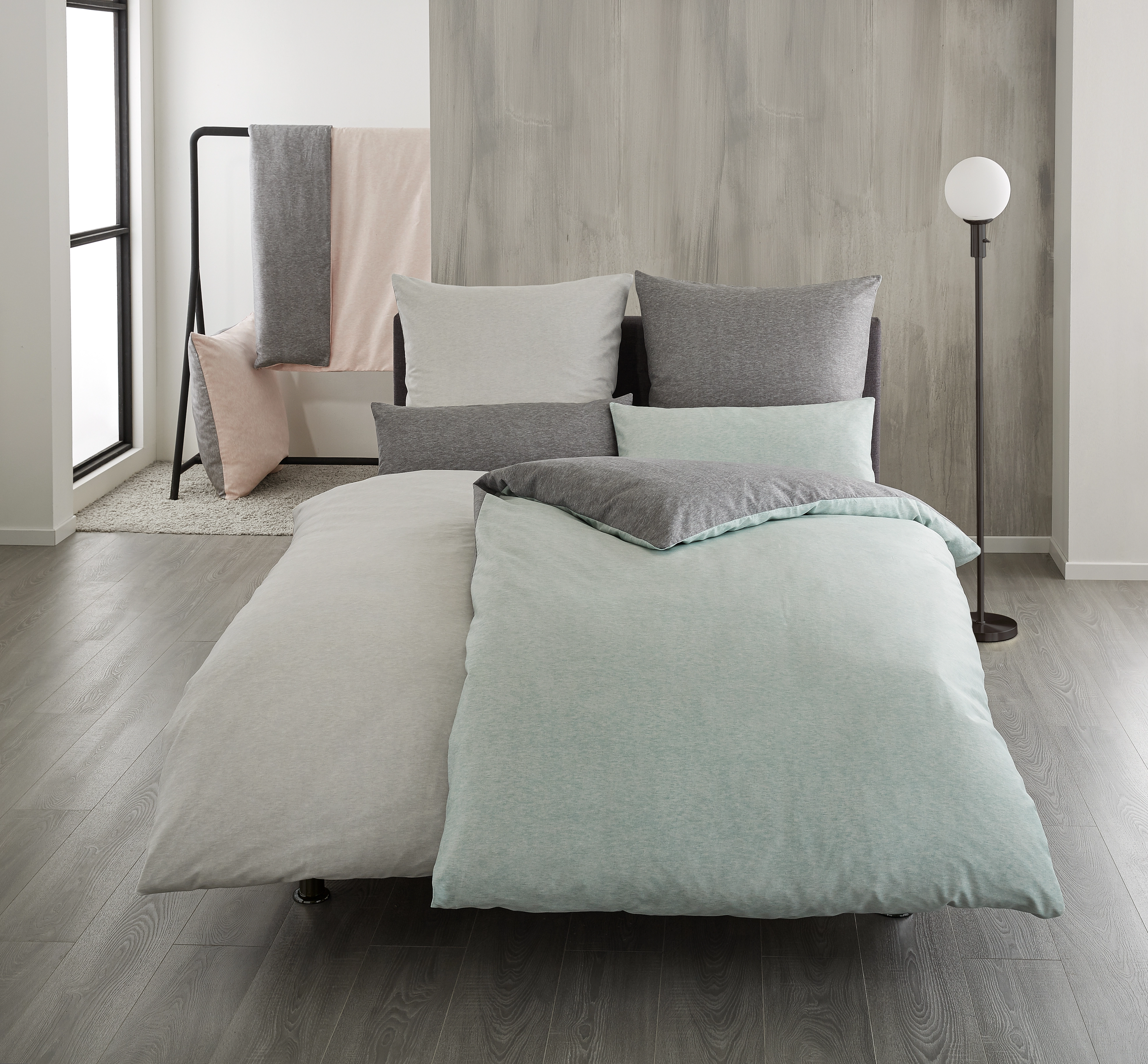 Bettwäsche-Set in sanften Pastelltönen, bestehend aus Bettbezug und Kissenbezügen, auf modernem Bett mit grauer Wand im Hintergrund.