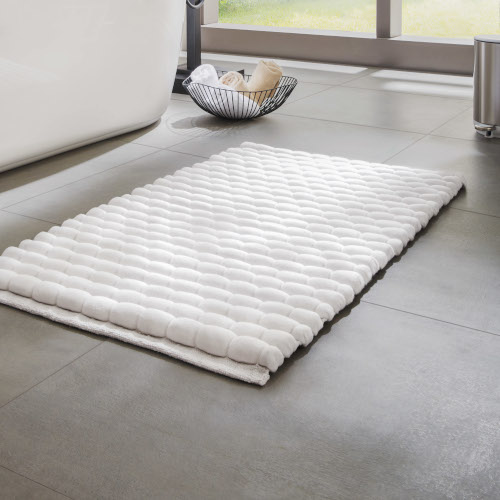 Weißer, gepolsterter Badezimmerteppich auf grauem Fliesenboden, im Hintergrund ein Fenster mit Blick ins Grüne.