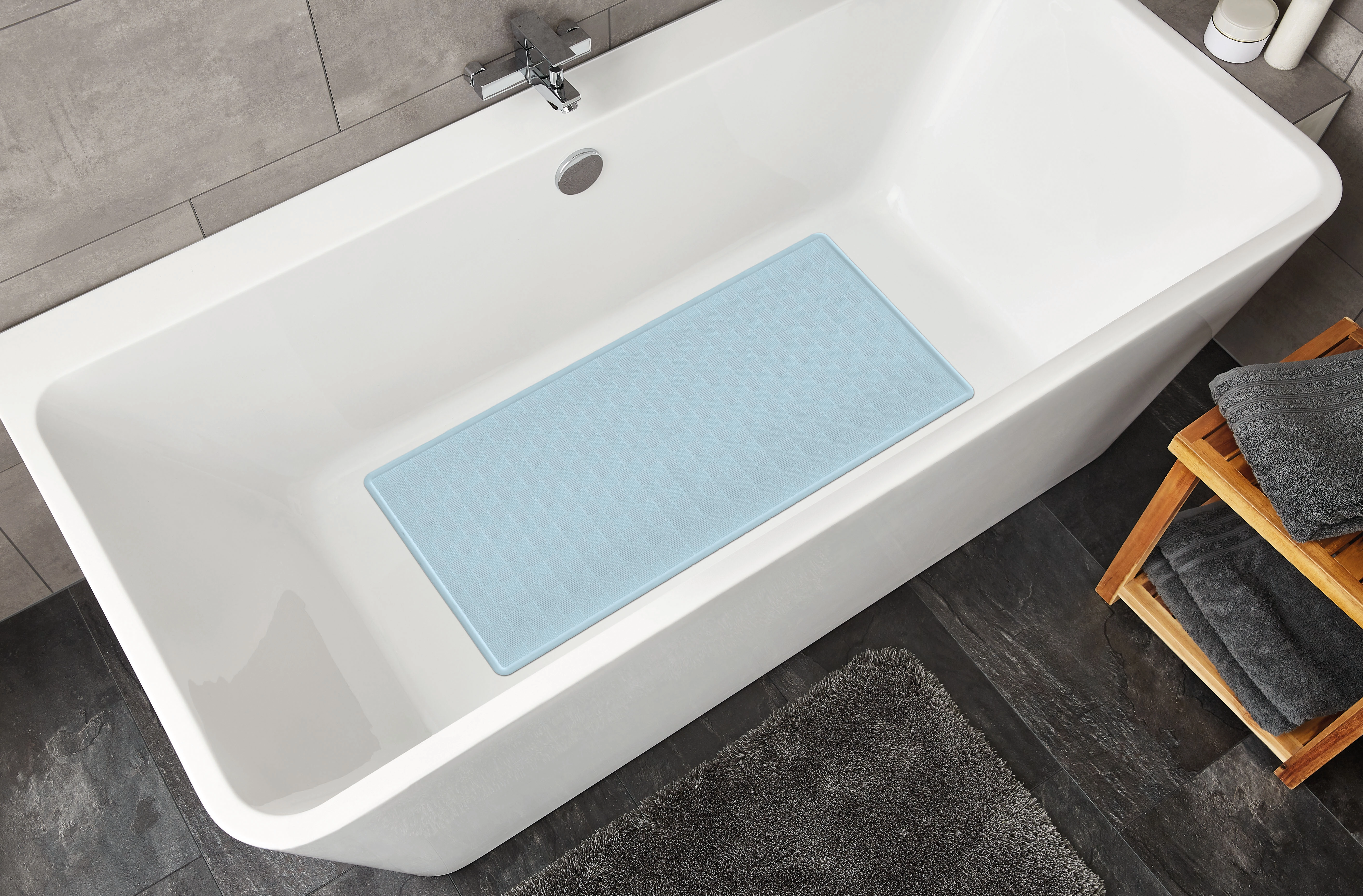Blaue Badematte in einer modernen weißen Badewanne, umgeben von grauen Fliesen und Handtüchern auf einem Holzregal.