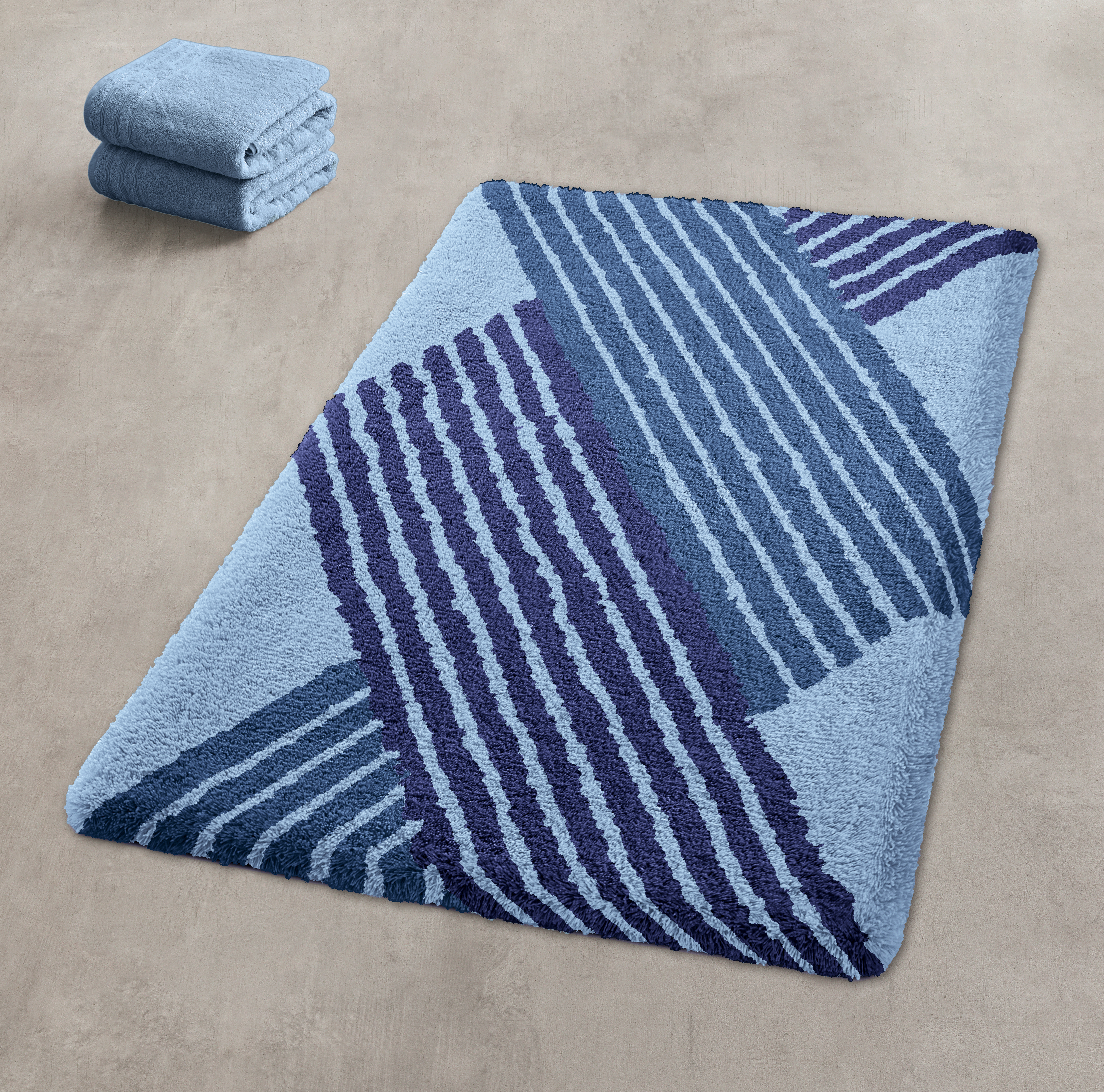 Blaue Badematte mit geometrischem Muster in verschiedenen Blautönen, daneben gefaltete Handtücher auf beigem Boden.