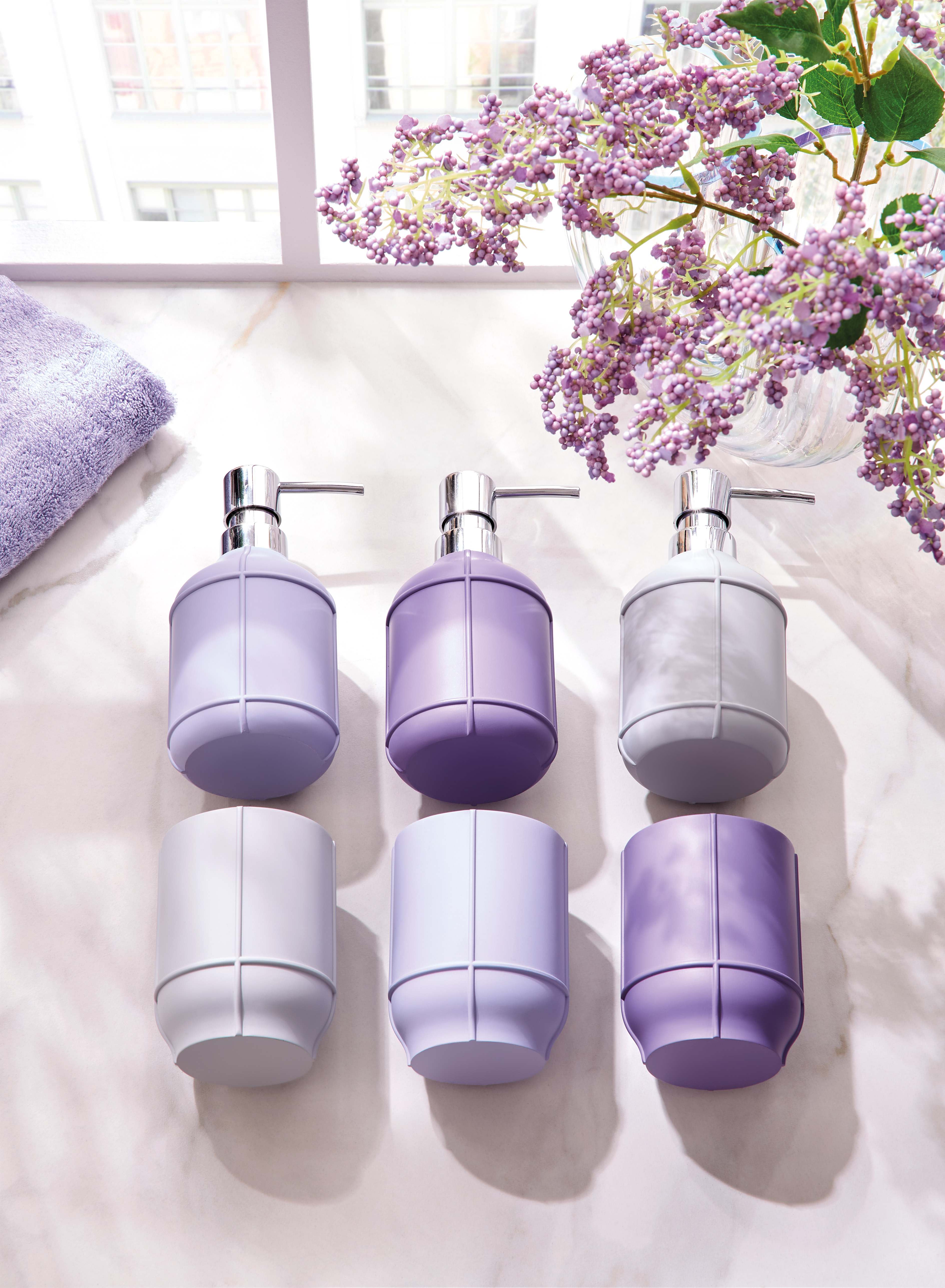 Seifenspender in verschiedenen Lilatönen auf einer Marmoroberfläche, dekoriert mit Fliederblüten im Hintergrund.