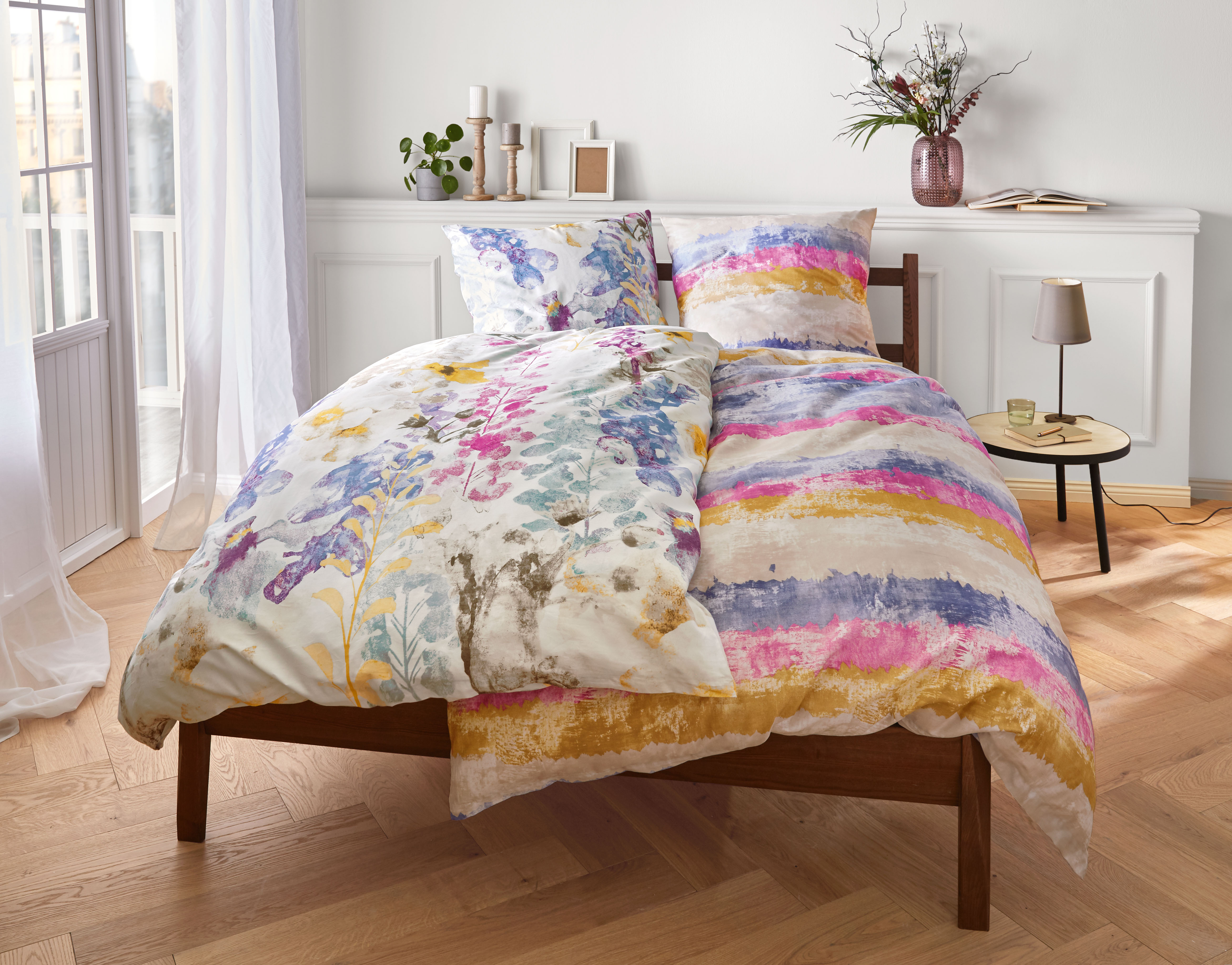 Bettwäsche-Set mit buntem Blumen- und Streifenmuster auf einem Holzrahmenbett in einem hellen, stilvollen Schlafzimmer.