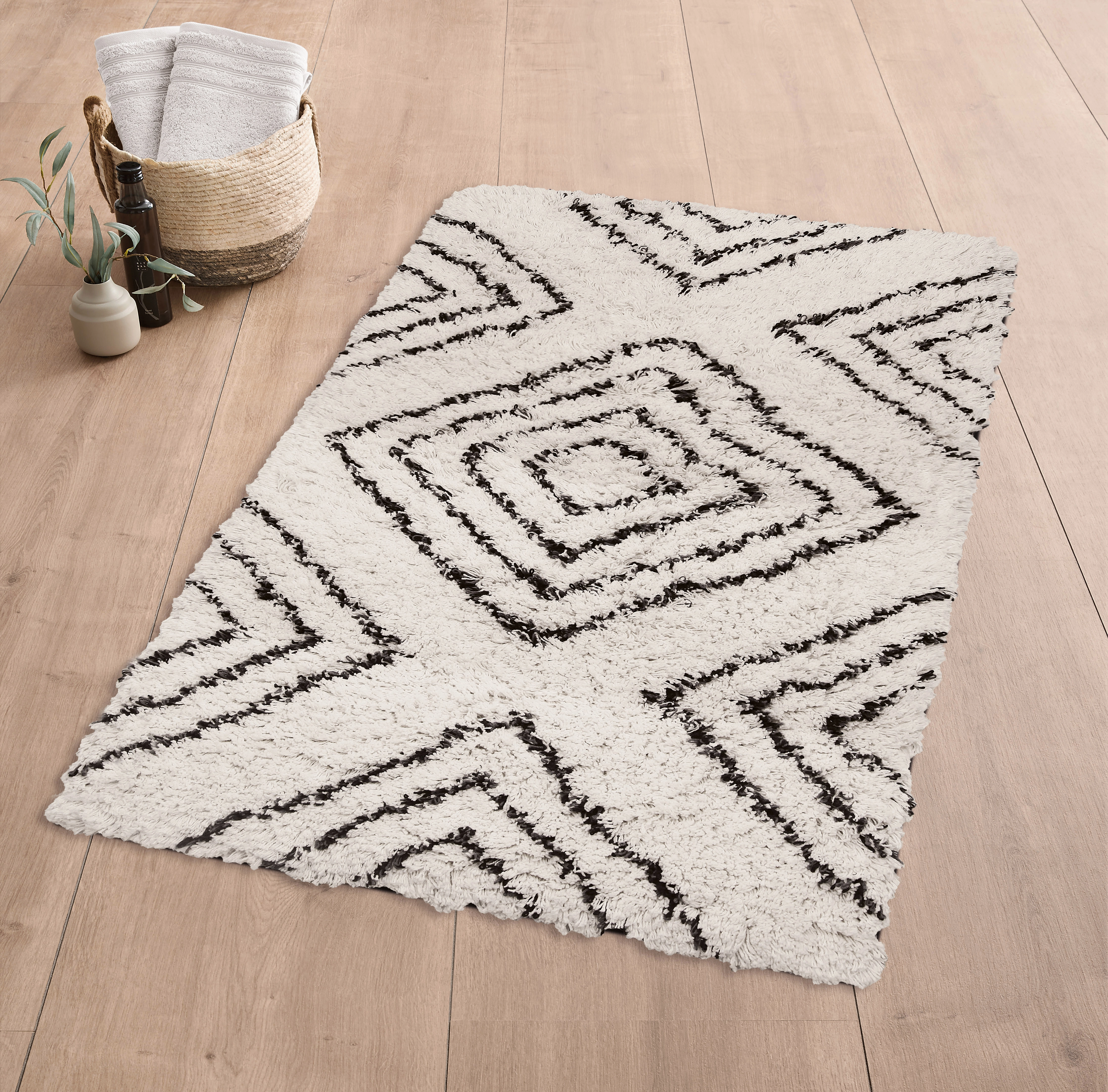 Moderner, cremefarbener Teppich mit schwarzem geometrischem Muster auf Holzfußboden, daneben ein Korb mit Handtüchern.