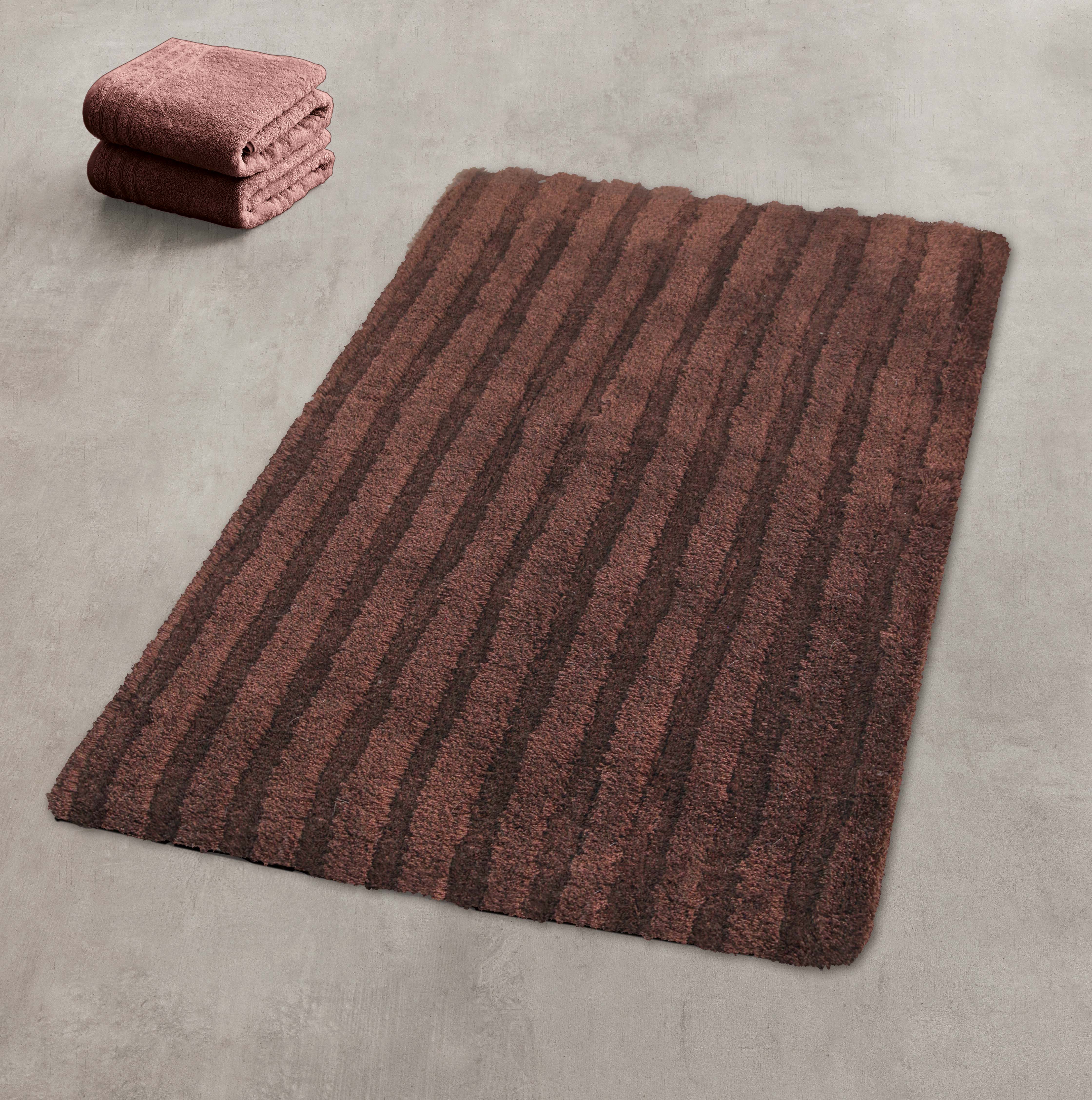 Brauner Badteppich mit Streifenmuster auf grauem Boden, daneben gefaltete Handtücher in passender Farbe.
