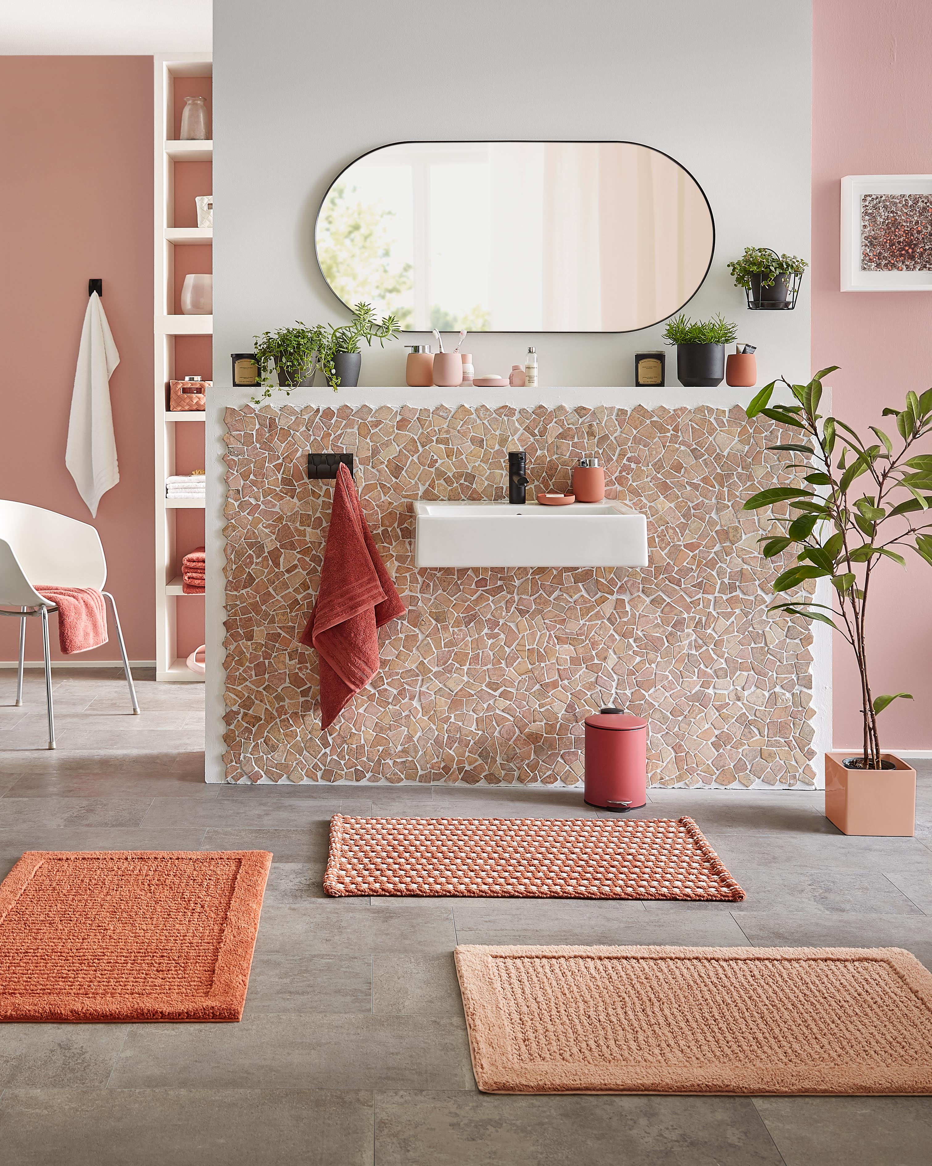 Modernes Badezimmer mit Mosaikfliesen, ovalem Spiegel, Pflanzen und orangefarbenen Badteppichen in verschiedenen Mustern.