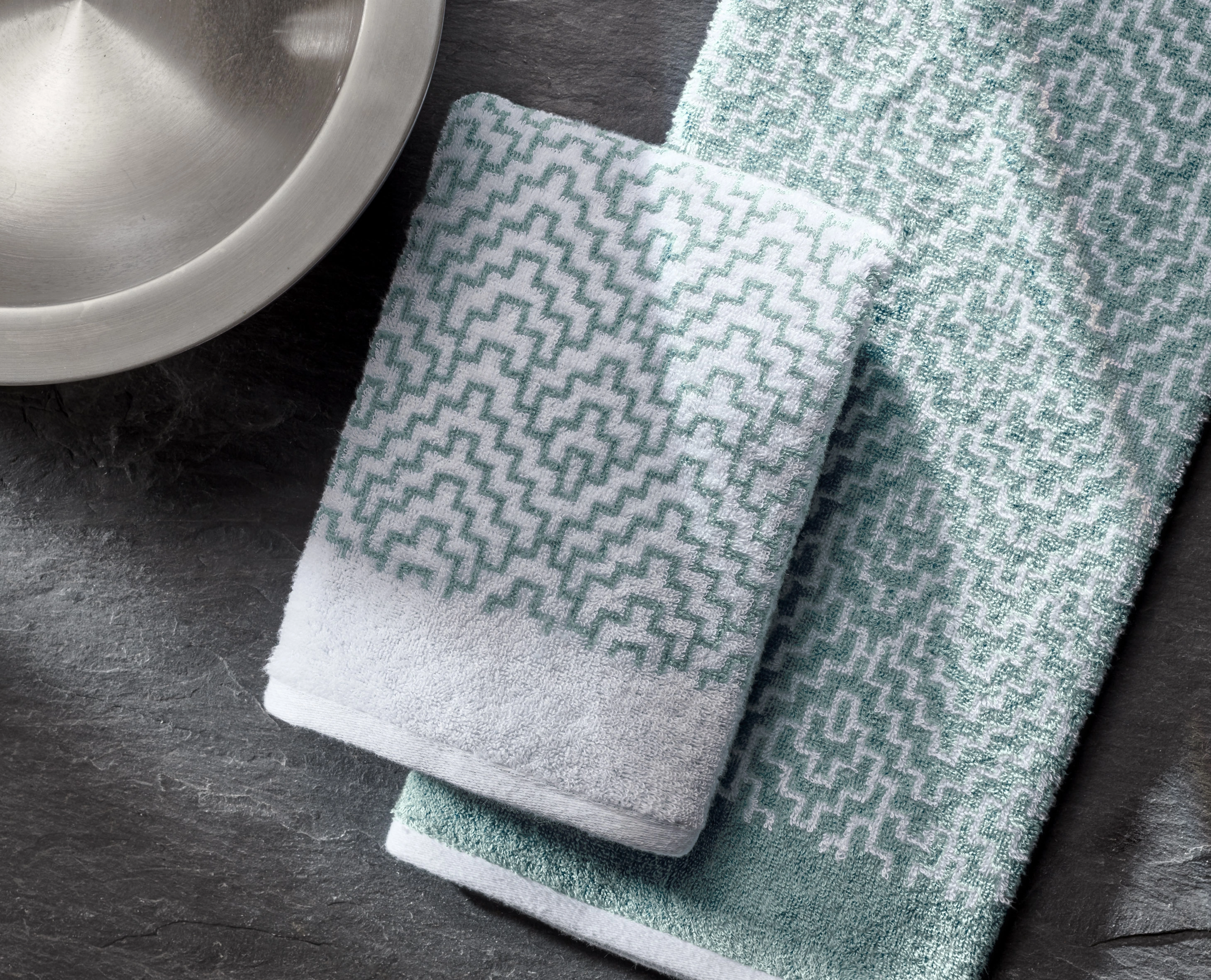 Handtuch-Set in Weiß und Mintgrün mit geometrischem Muster, auf dunkler Steinoberfläche arrangiert.