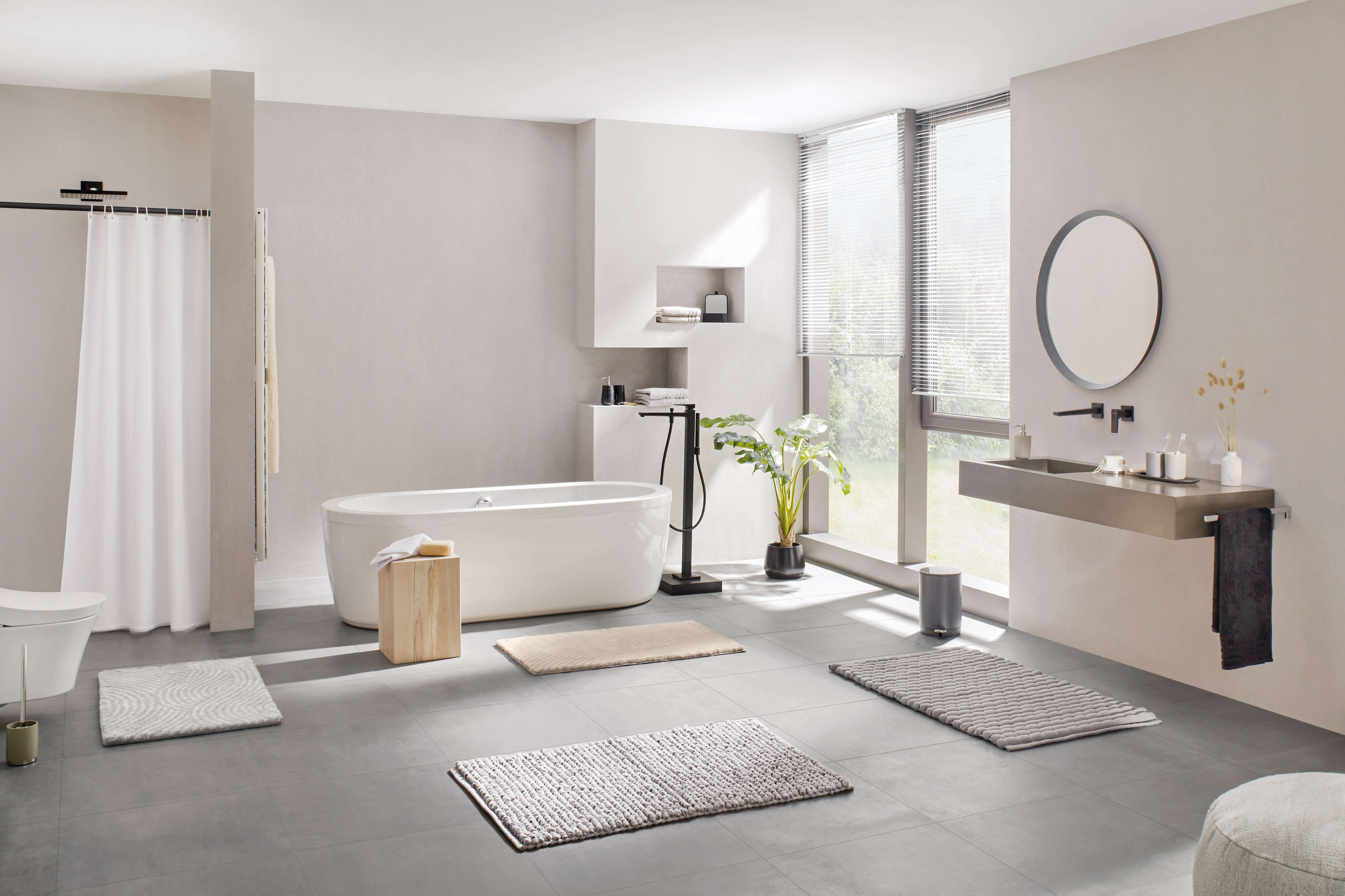Modernes Badezimmer mit freistehender Badewanne, Waschbecken, Spiegel und verschiedenen Badteppichen auf grauem Boden.
