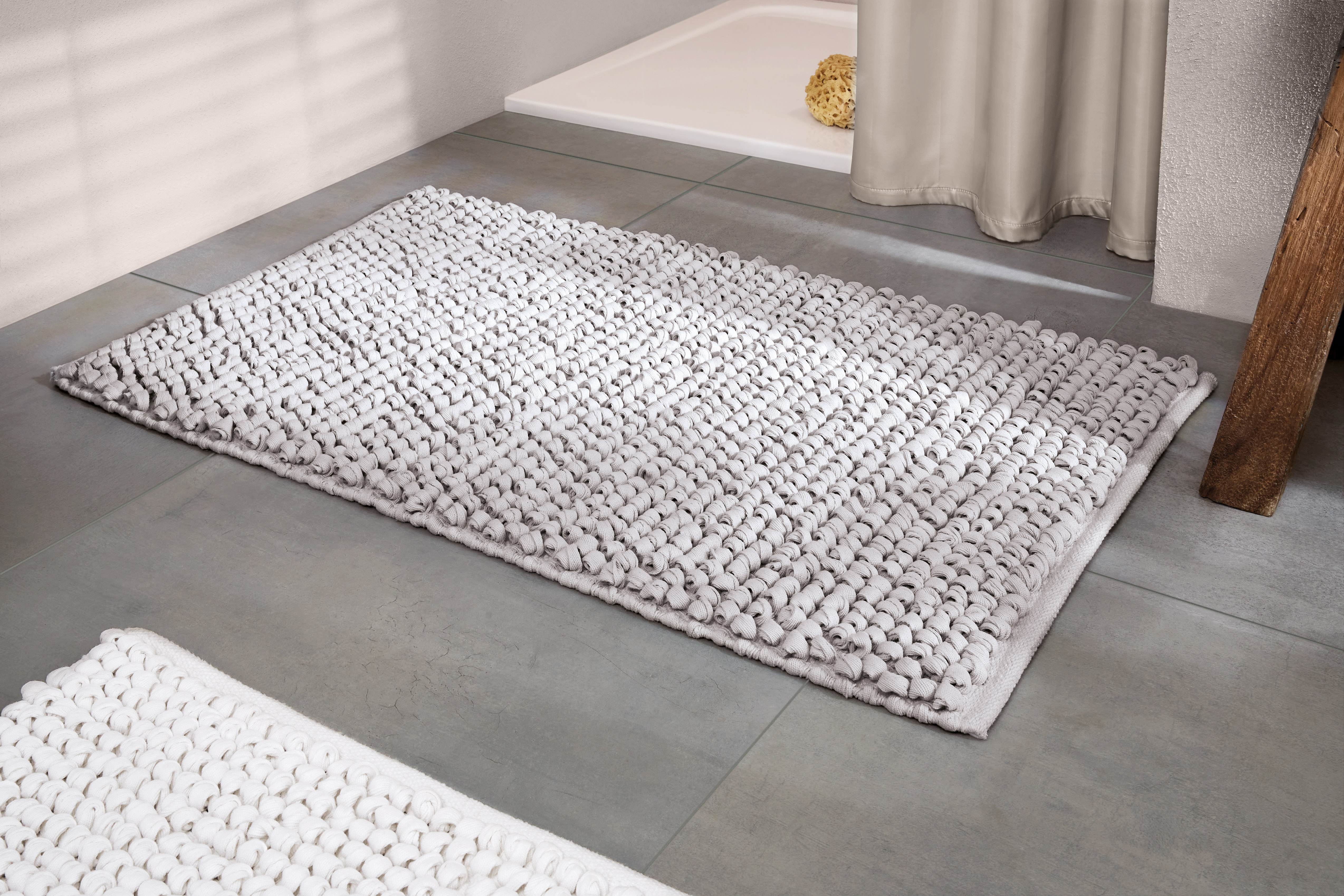 Weißer, strukturierter Badezimmerteppich auf grauem Fliesenboden, neben einer Dusche mit beigem Vorhang und einem Holzregal.