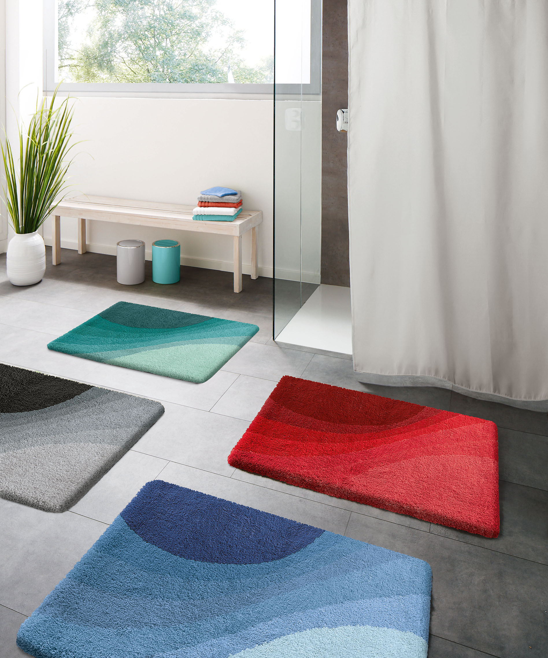 Bunte Badematten in verschiedenen Farben und Mustern auf grauem Badezimmerboden, mit Dusche, Bank und Pflanzen im Hintergrund.