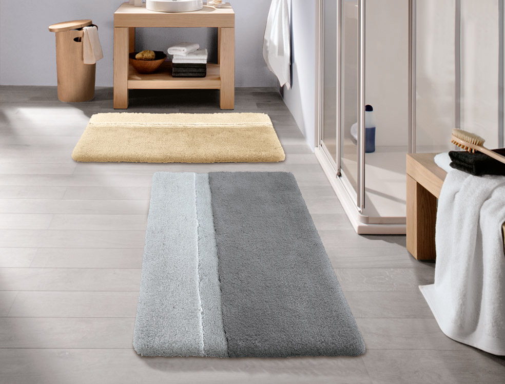 Badezimmer mit zwei flauschigen Badematten in Beige und Grau, moderne Einrichtung mit Holz- und Glaselementen.