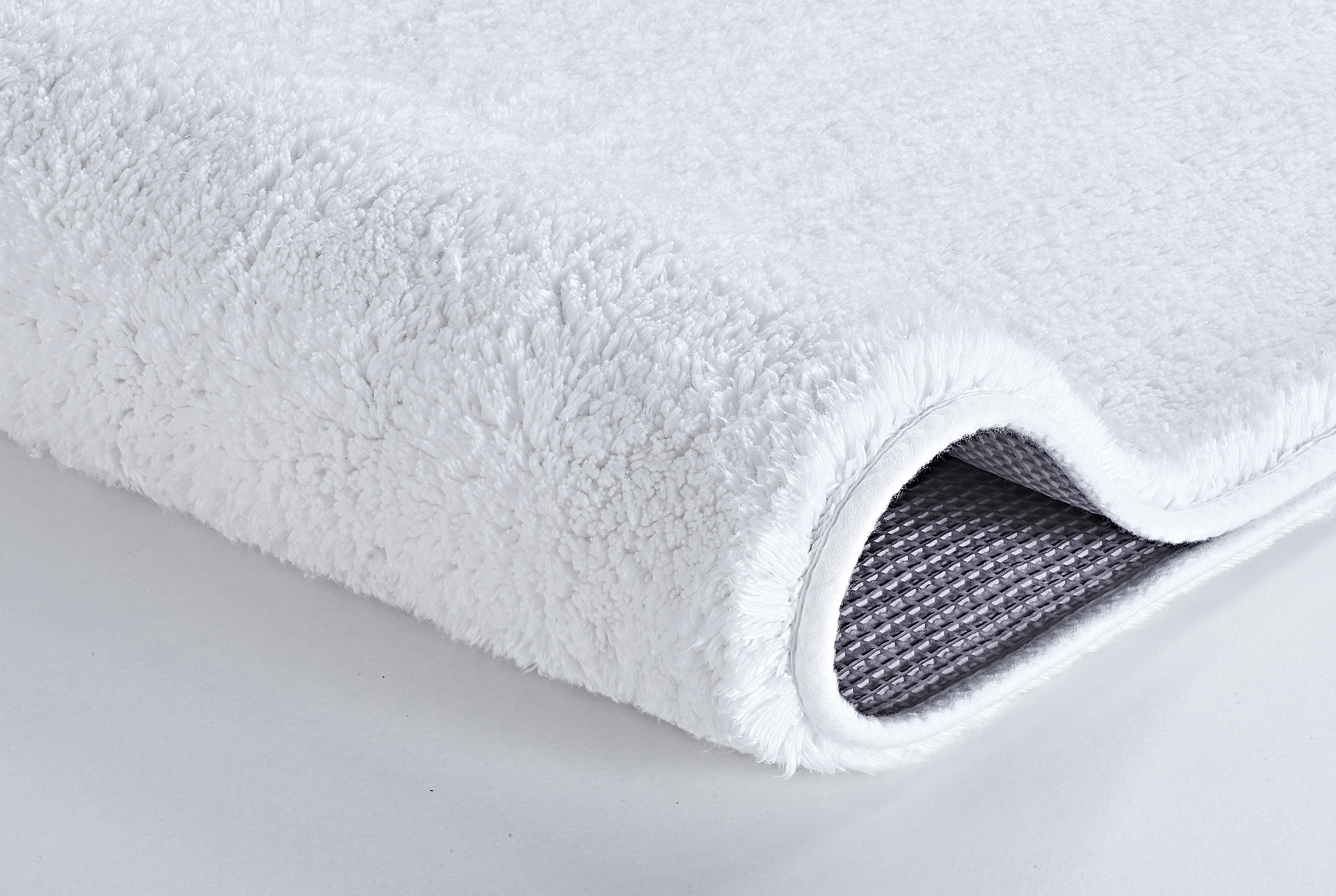 Weißer, flauschiger Badteppich mit rutschfester Unterseite in Nahaufnahme, zeigt die weiche Textur und die graue Rückseite.