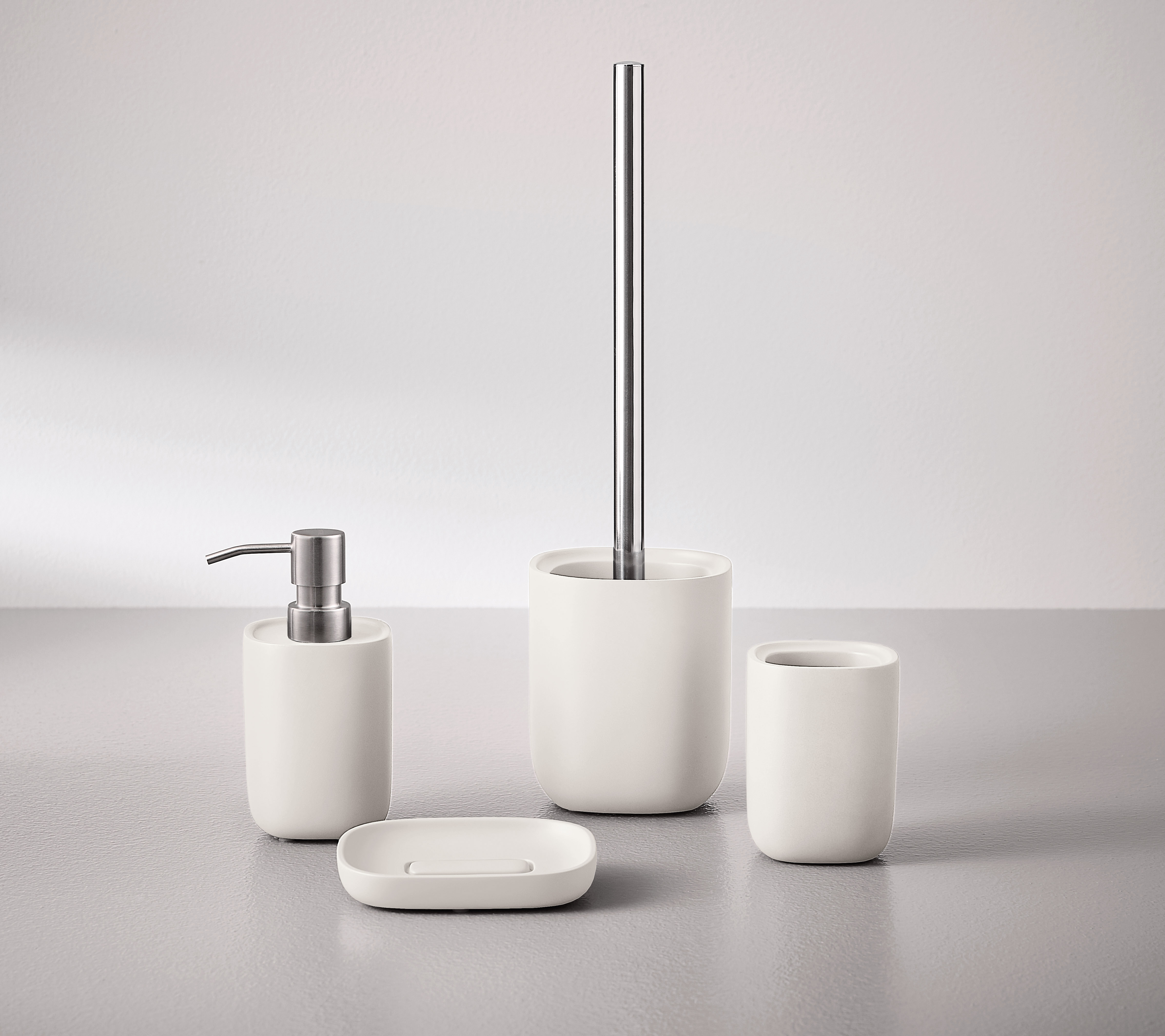 Weißes Badezimmer-Set aus Keramik mit Seifenspender, Zahnbürstenhalter, Seifenschale und WC-Bürste auf grauem Hintergrund.