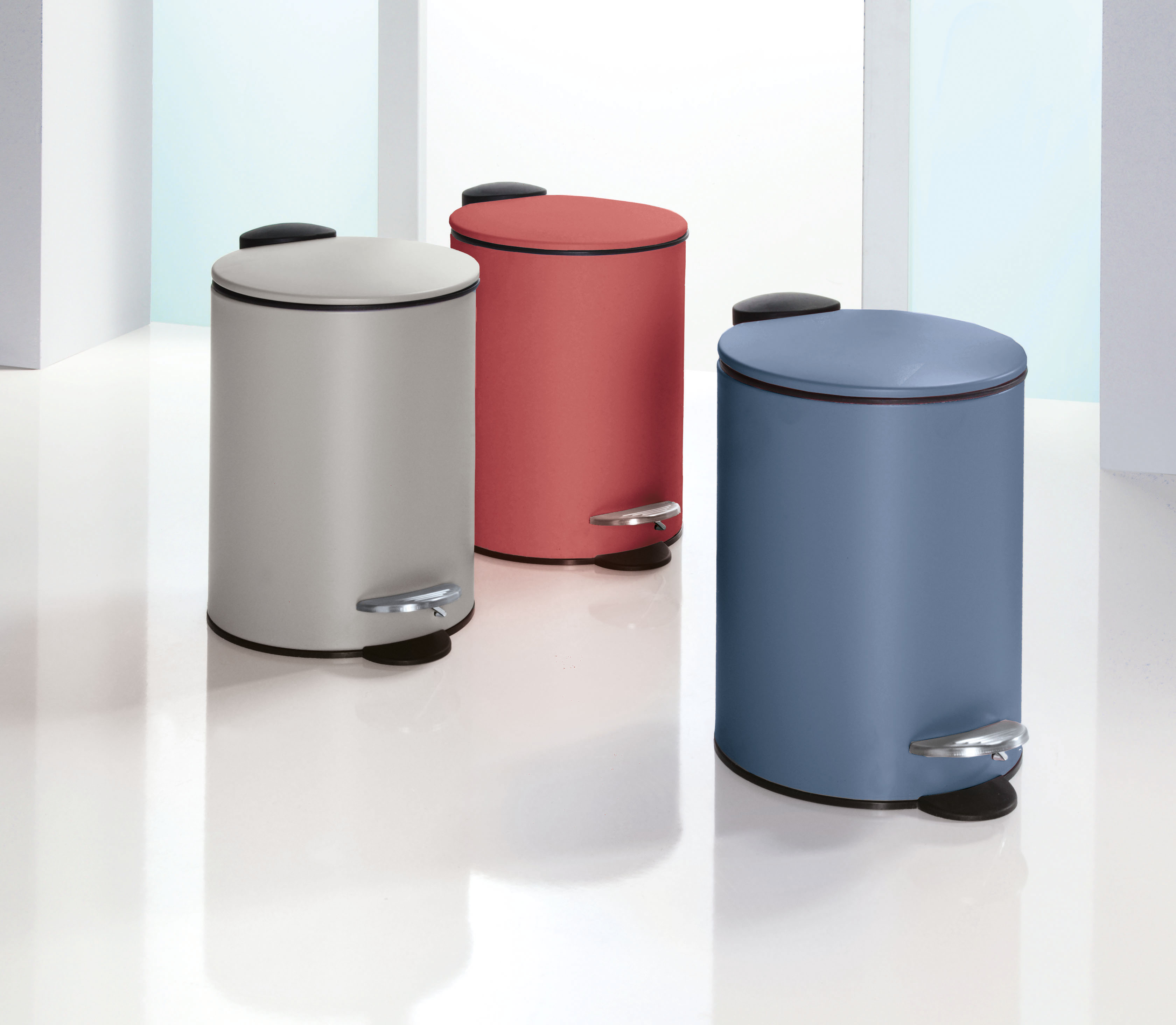 Drei runde Tretmülleimer in Grau, Rot und Blau mit glänzenden Pedalen auf glänzendem Boden.