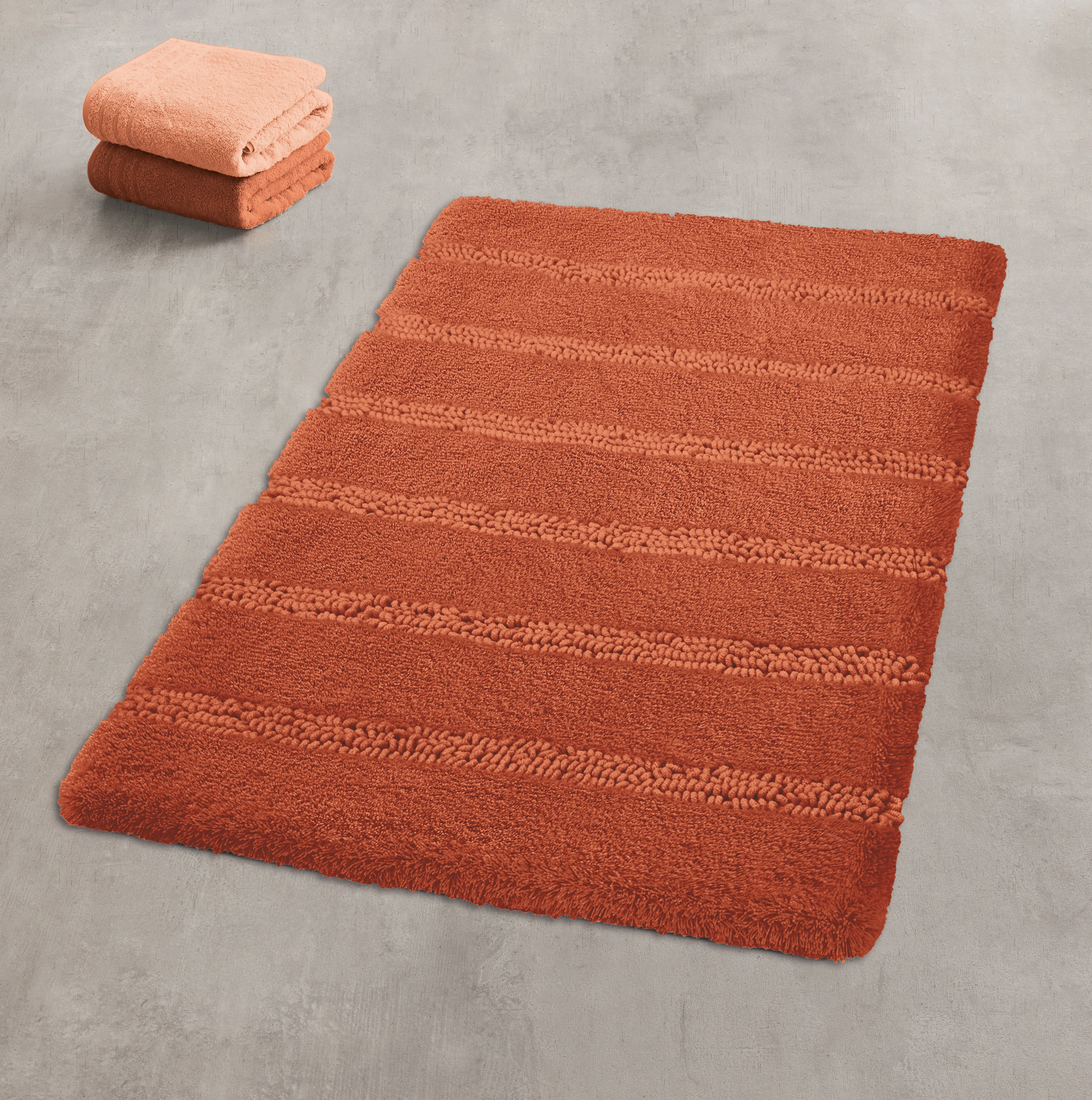 Roter Badteppich mit Streifenmuster auf grauem Boden, daneben gefaltete Handtücher.