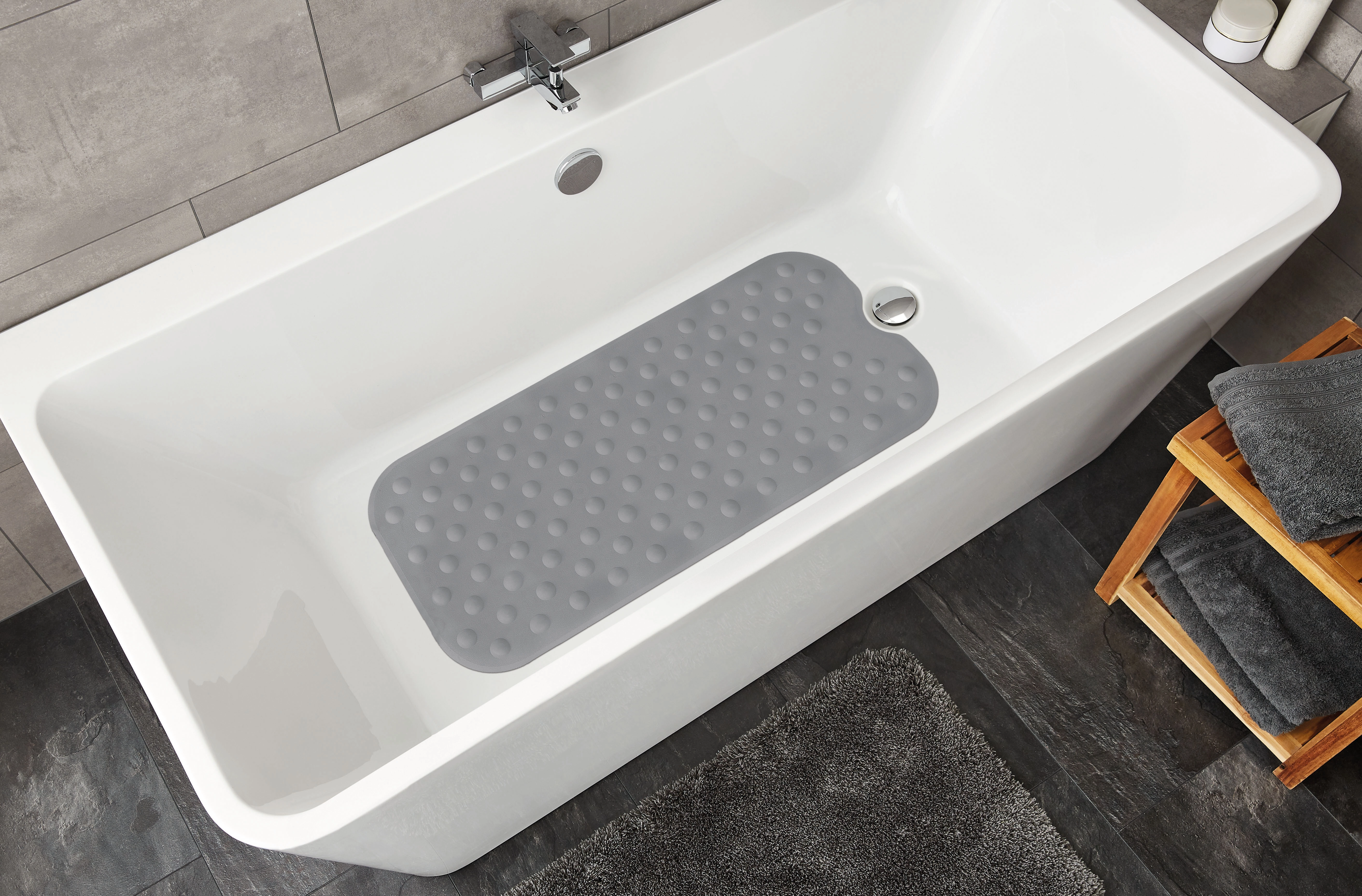 Graue Badewannenmatte mit rutschfester Oberfläche in einer modernen weißen Badewanne, umgeben von grauen Fliesen und Handtüchern.