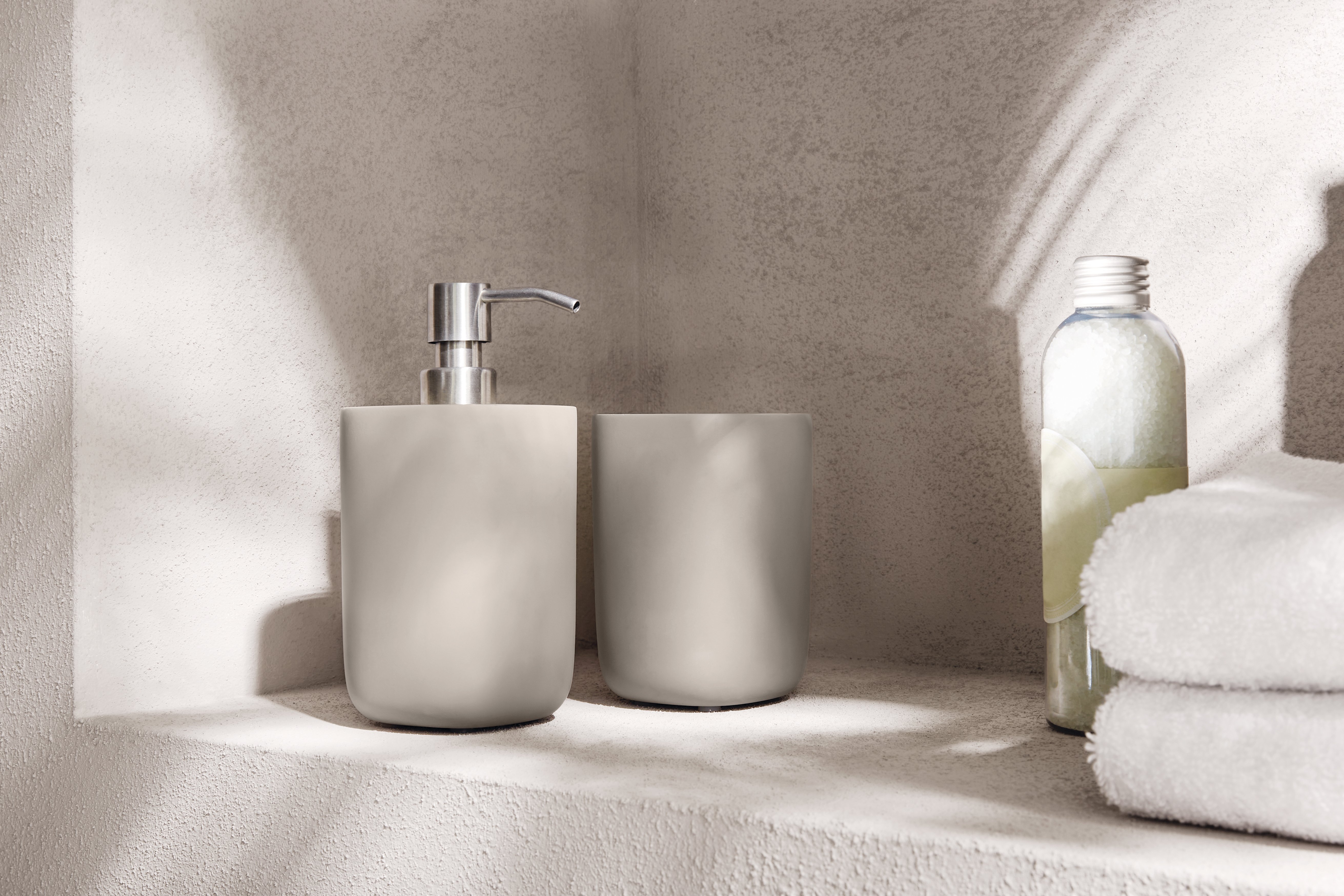 Elegantes Badezimmer-Set in Beige mit Seifenspender und Becher, daneben weiße Handtücher und eine Flasche.