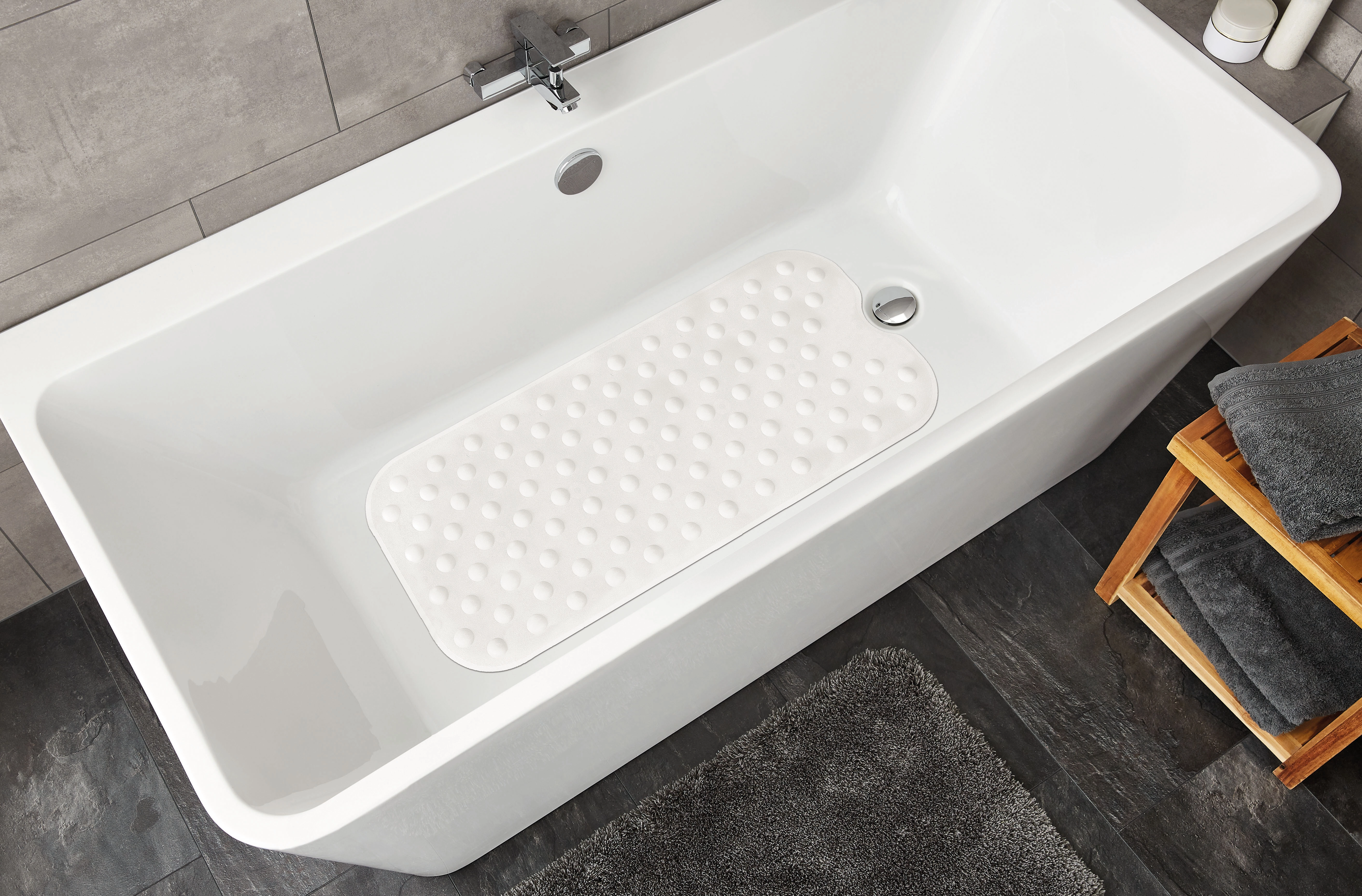 Weiße Badewannenmatte mit rutschfester Oberfläche in einer modernen Badewanne, umgeben von grauen Fliesen und Handtüchern.