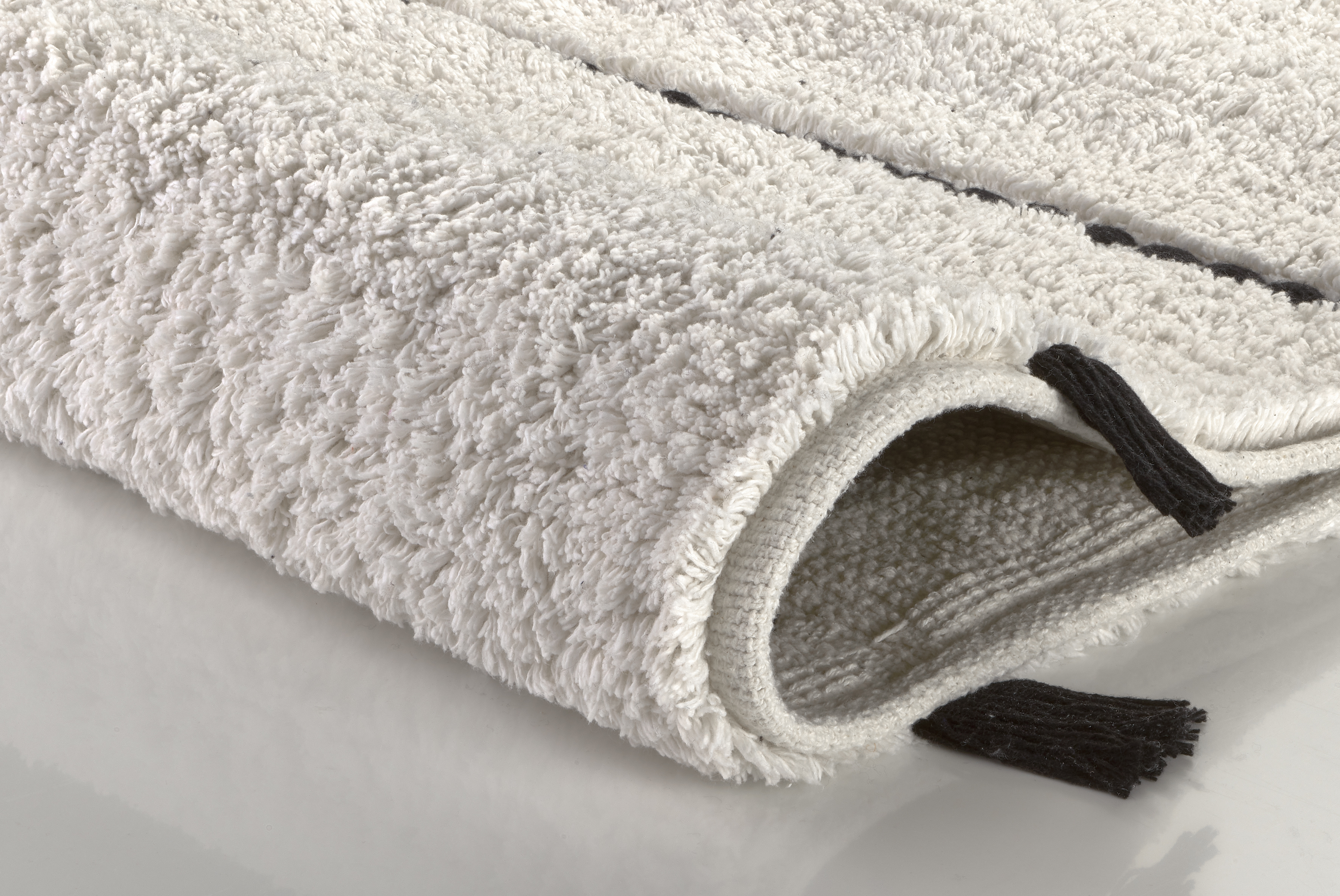 Weißer, flauschiger Badteppich mit schwarzem Randdetail, aufgerollt und auf einer glatten Oberfläche liegend.