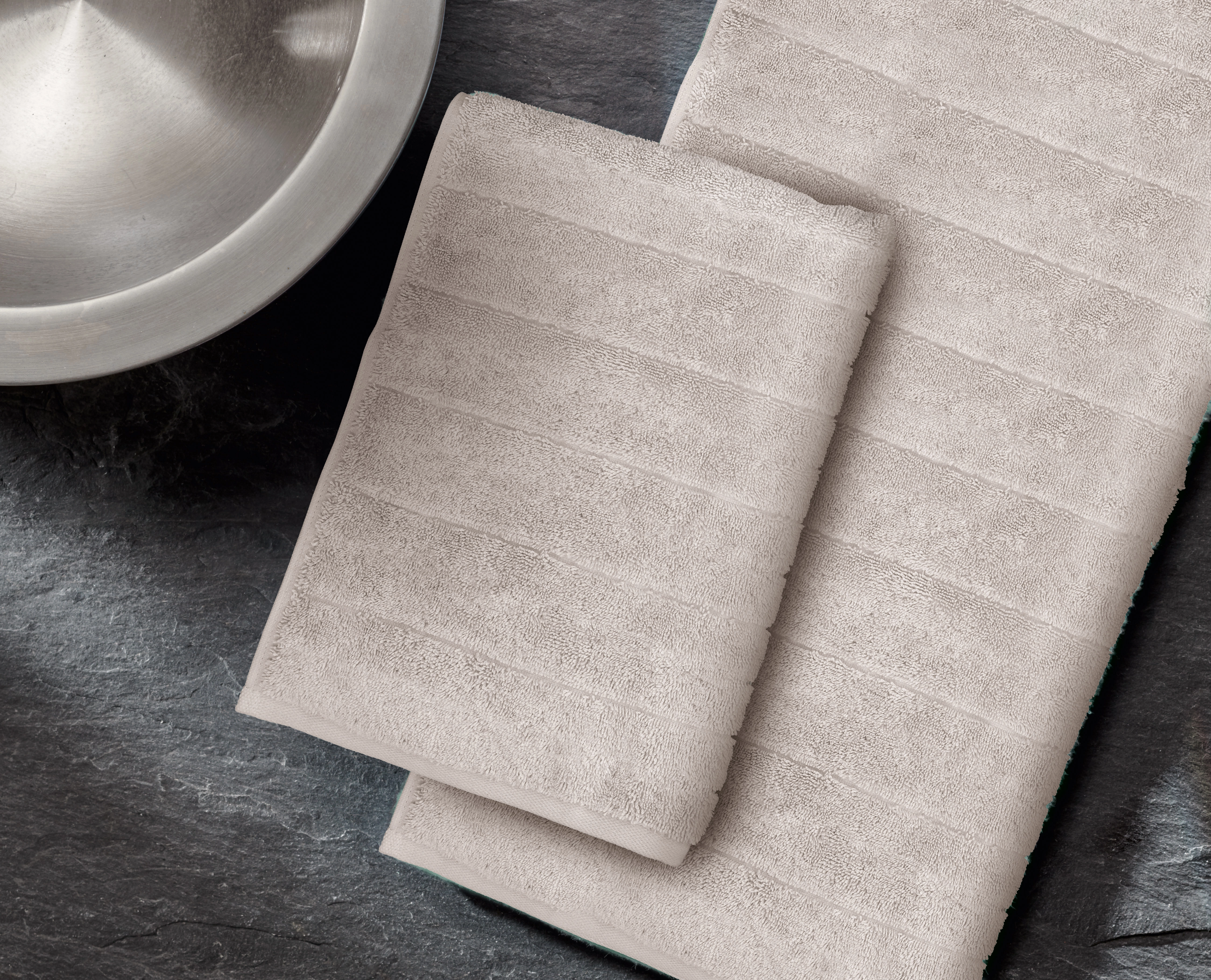 Beige Handtücher auf dunklem Untergrund, elegant gefaltet, mit feiner Streifenstruktur, neben einer silbernen Schale.