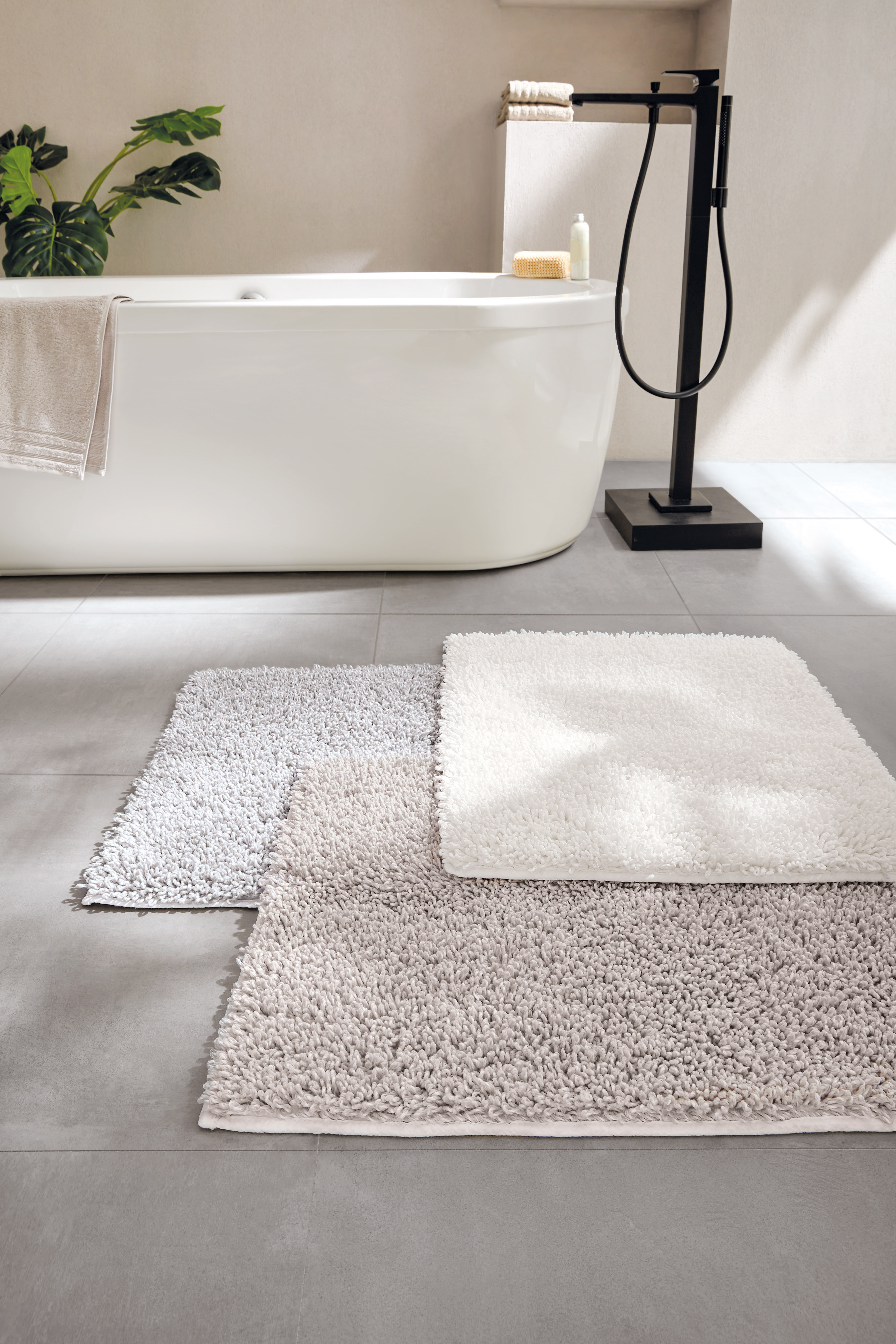 Flauschige Badematten in Weiß, Grau und Beige vor einer modernen Badewanne mit schwarzer Armatur.
