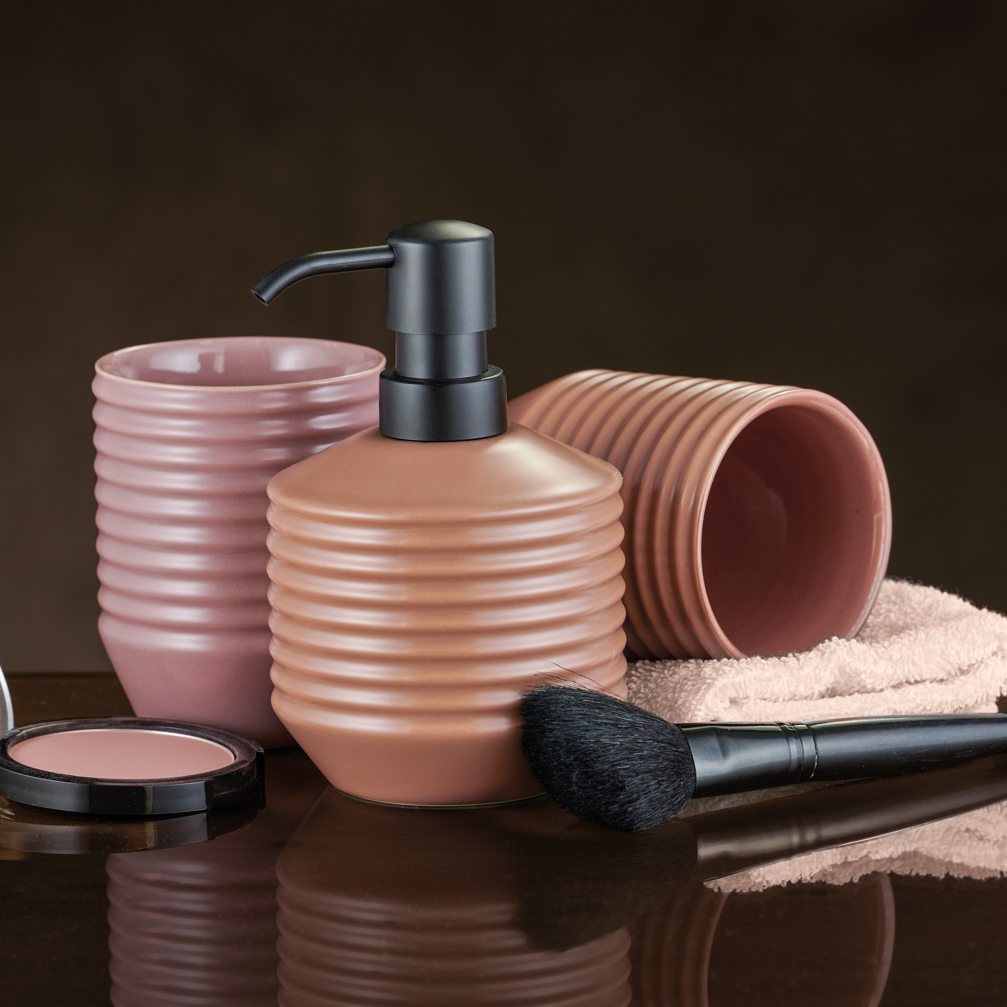 Elegantes Badezimmer-Set in Rosa mit Seifenspender, Bechern und Schminkpinsel auf einem Handtuch.