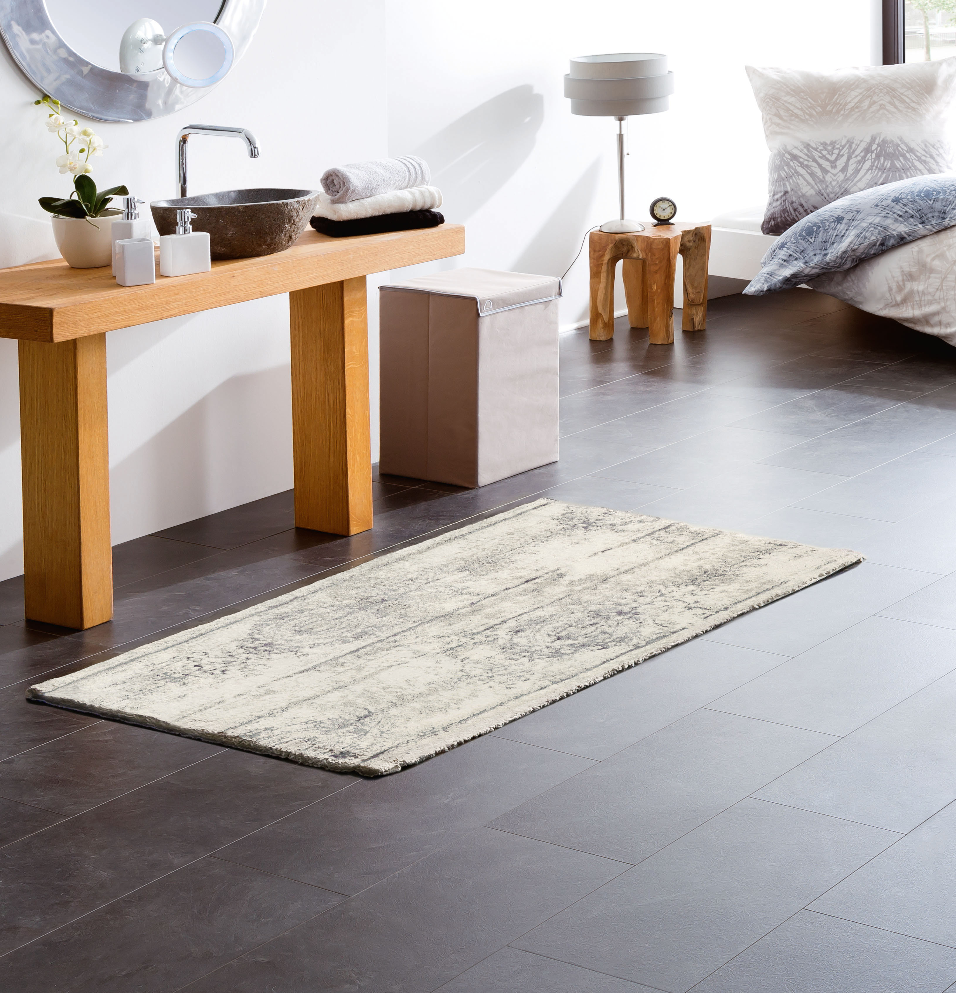 Moderner Badezimmerteppich in Beige und Grau, platziert auf dunklem Fliesenboden, neben einem Holzwaschtisch und einem Bett.