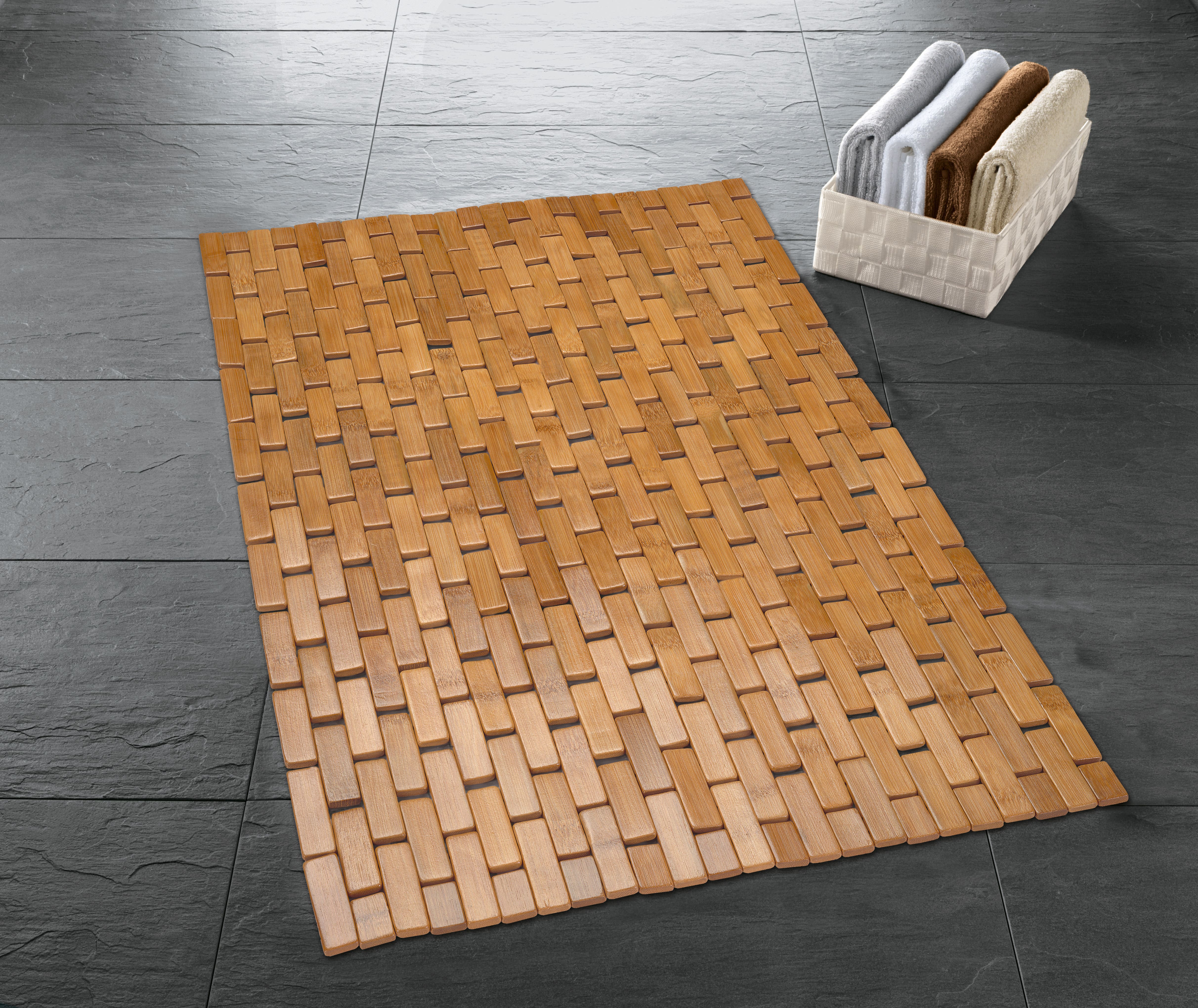 Holz-Badematte auf dunklem Fliesenboden, daneben ein Korb mit gefalteten Handtüchern in verschiedenen Farben.