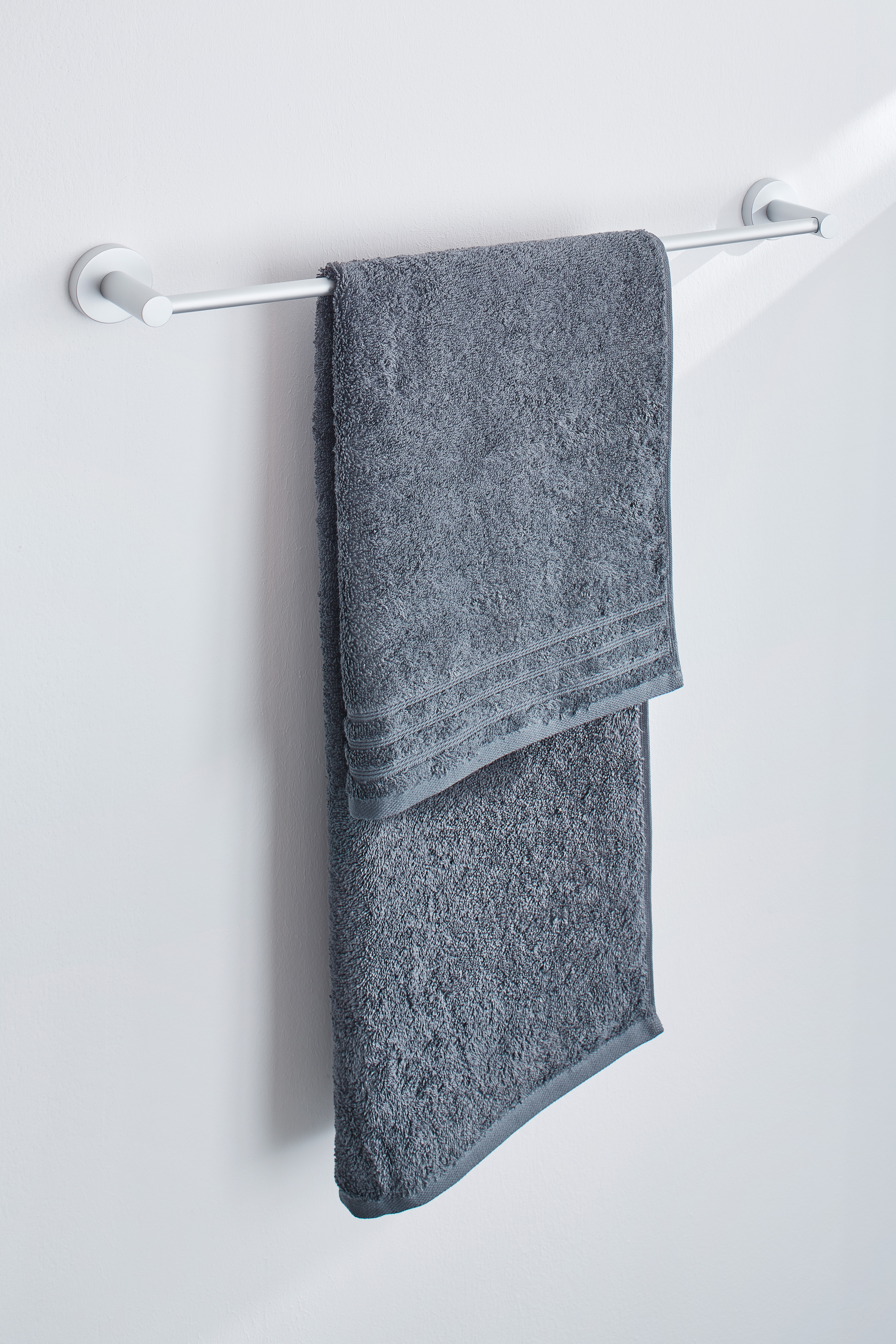 Graues Handtuch aus weicher Baumwolle, aufgehängt an einem weißen Handtuchhalter vor einer weißen Wand.