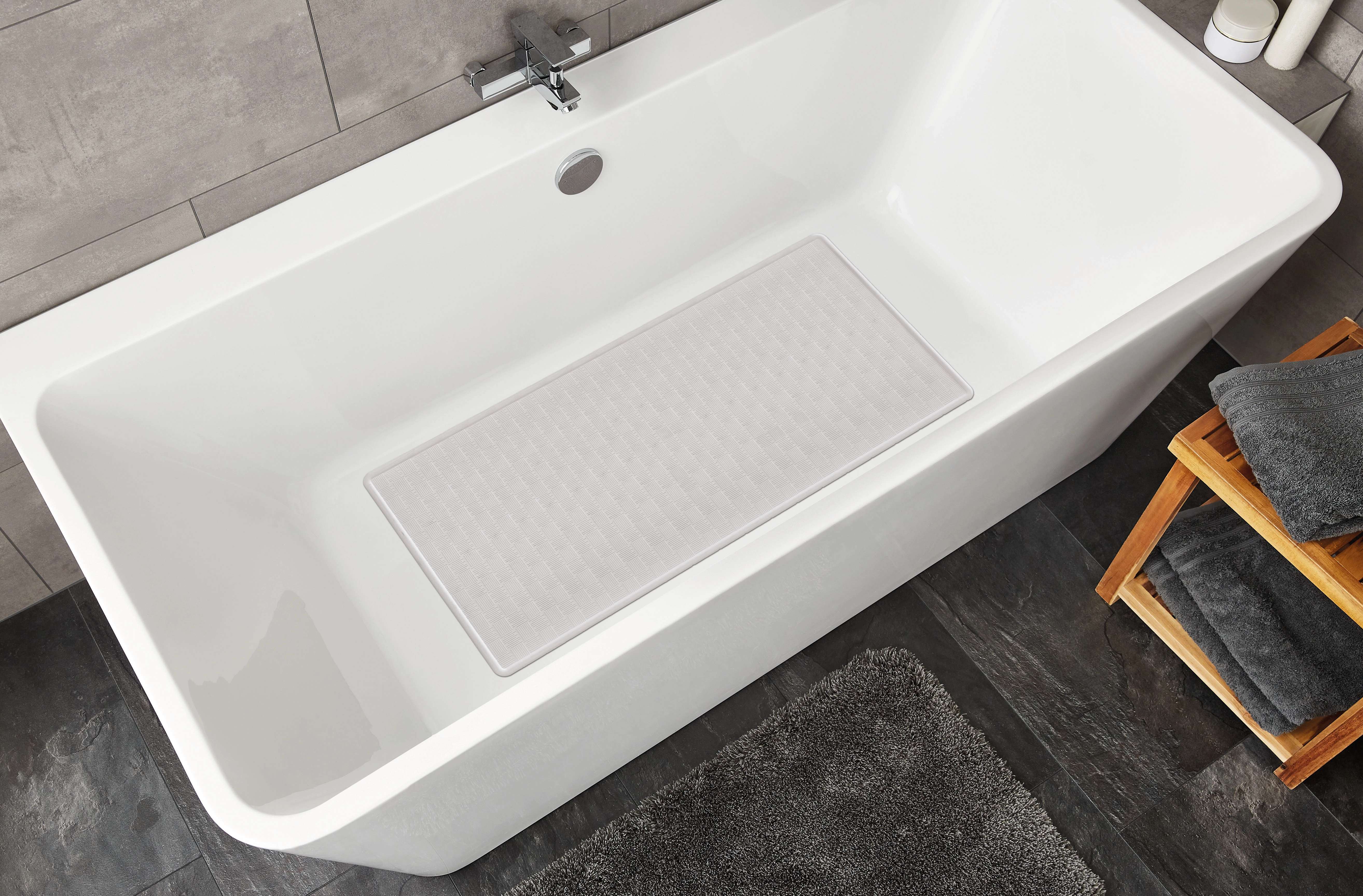 Weiße Badewanne mit rechteckiger, hellgrauer Badematte, umgeben von grauen Fliesen und Handtüchern auf einem Holzregal.