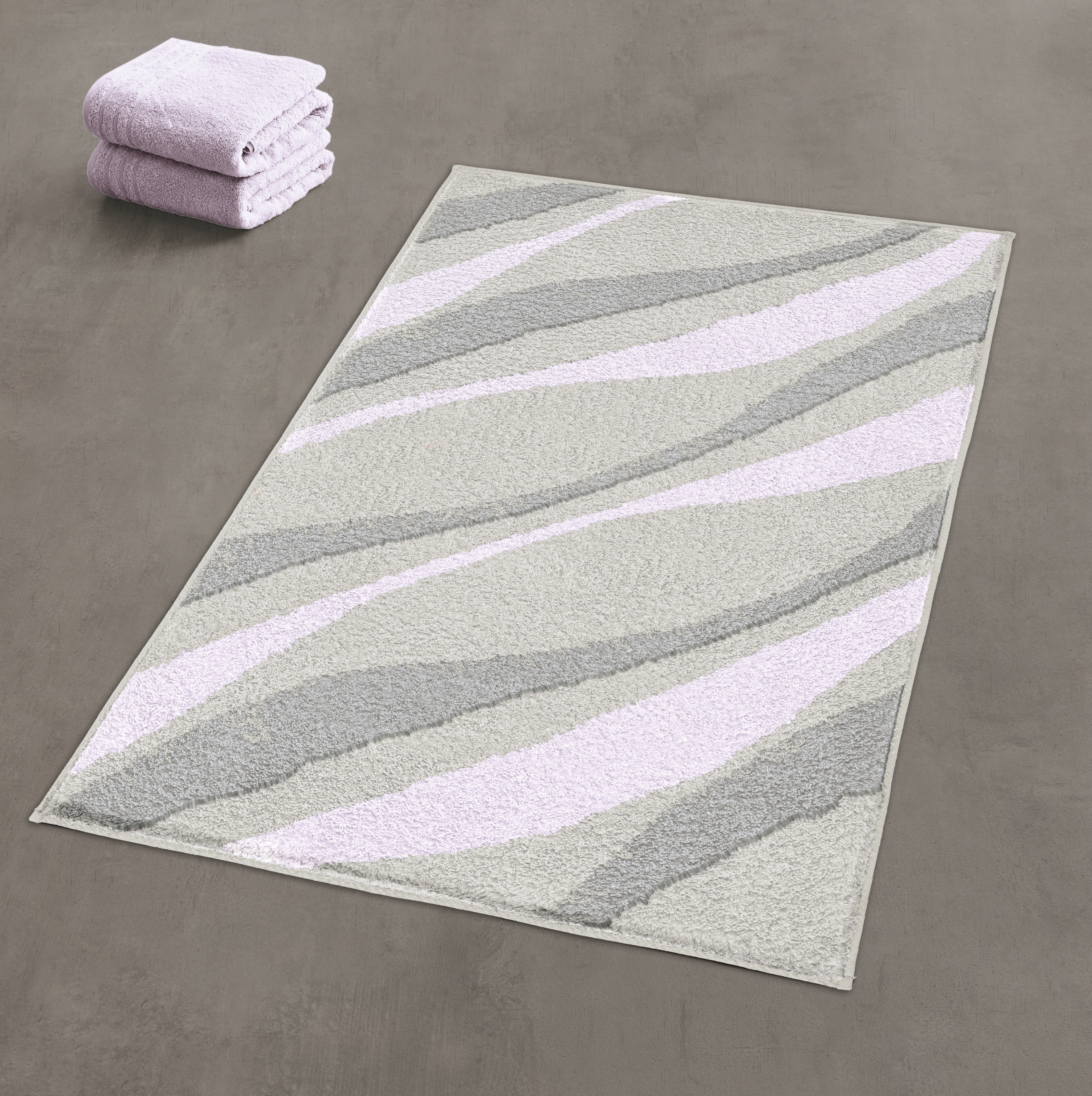 Moderner Badezimmerteppich mit wellenförmigem Design in Grau- und Lilatönen, daneben gefaltete Handtücher.